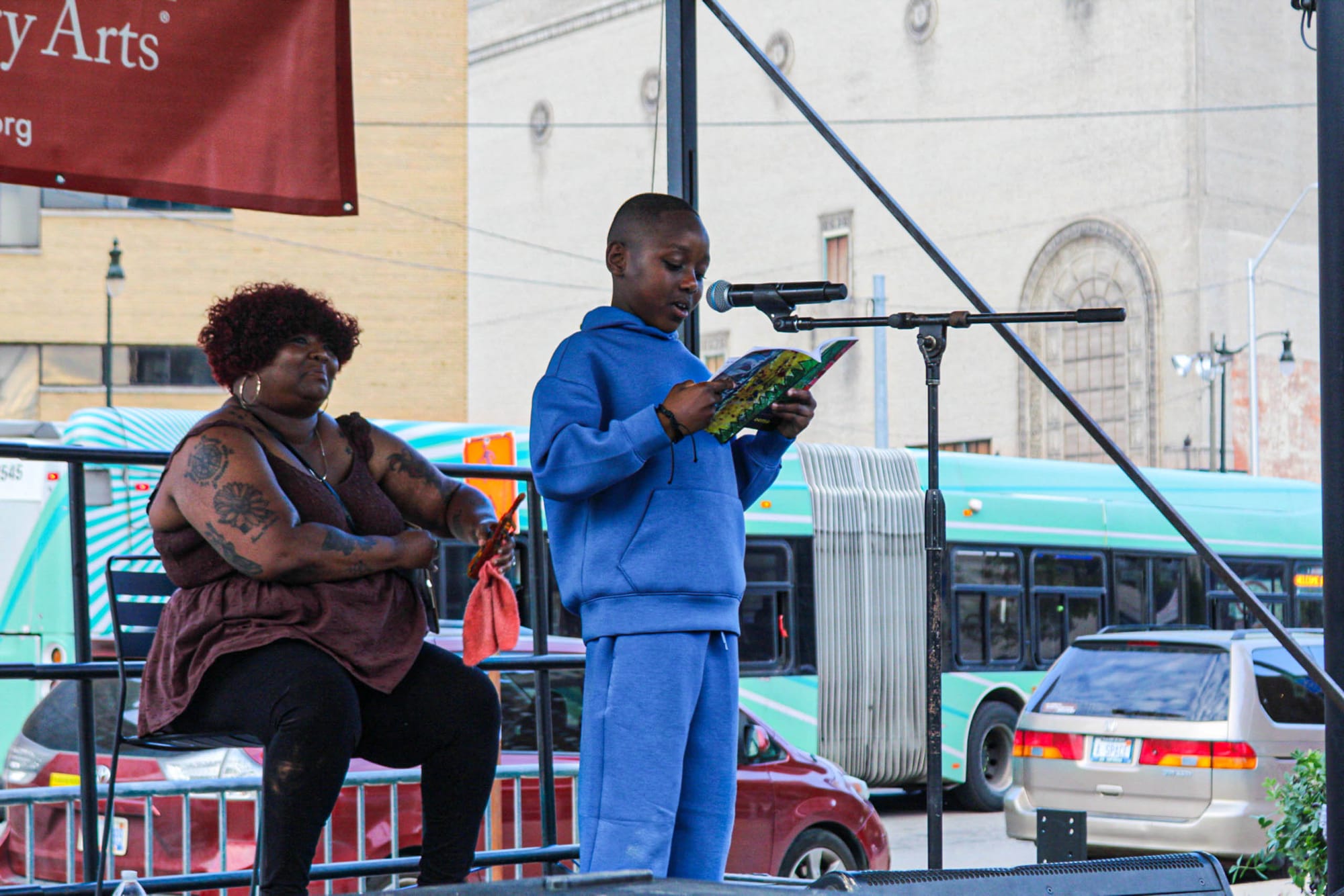 A child is on stage with one adult behind him as he reads a poem from his notebook into a microphone.