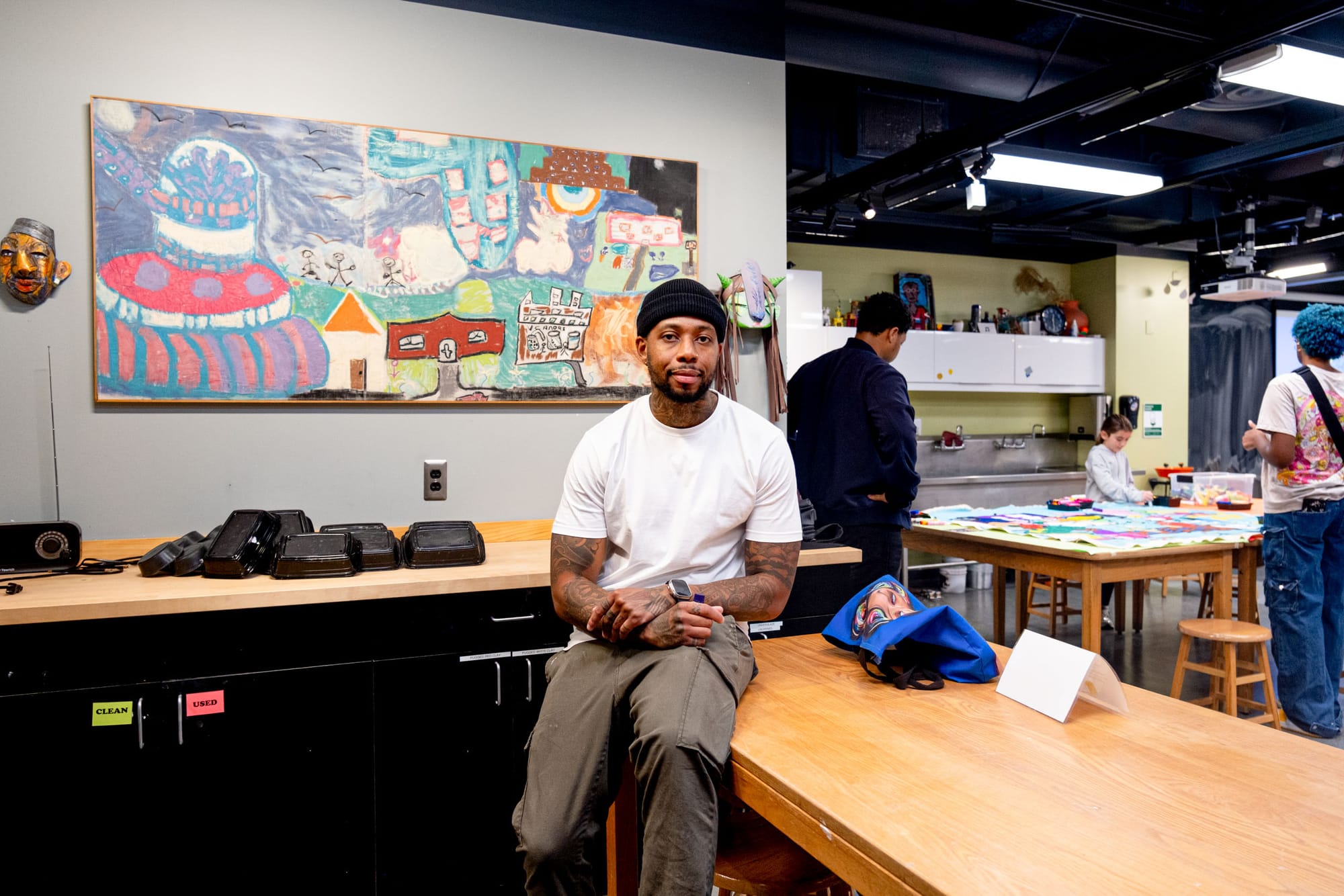 A man wearing a white shirt and black beanie sits on a wooden desk while people in the background engage with artwork.