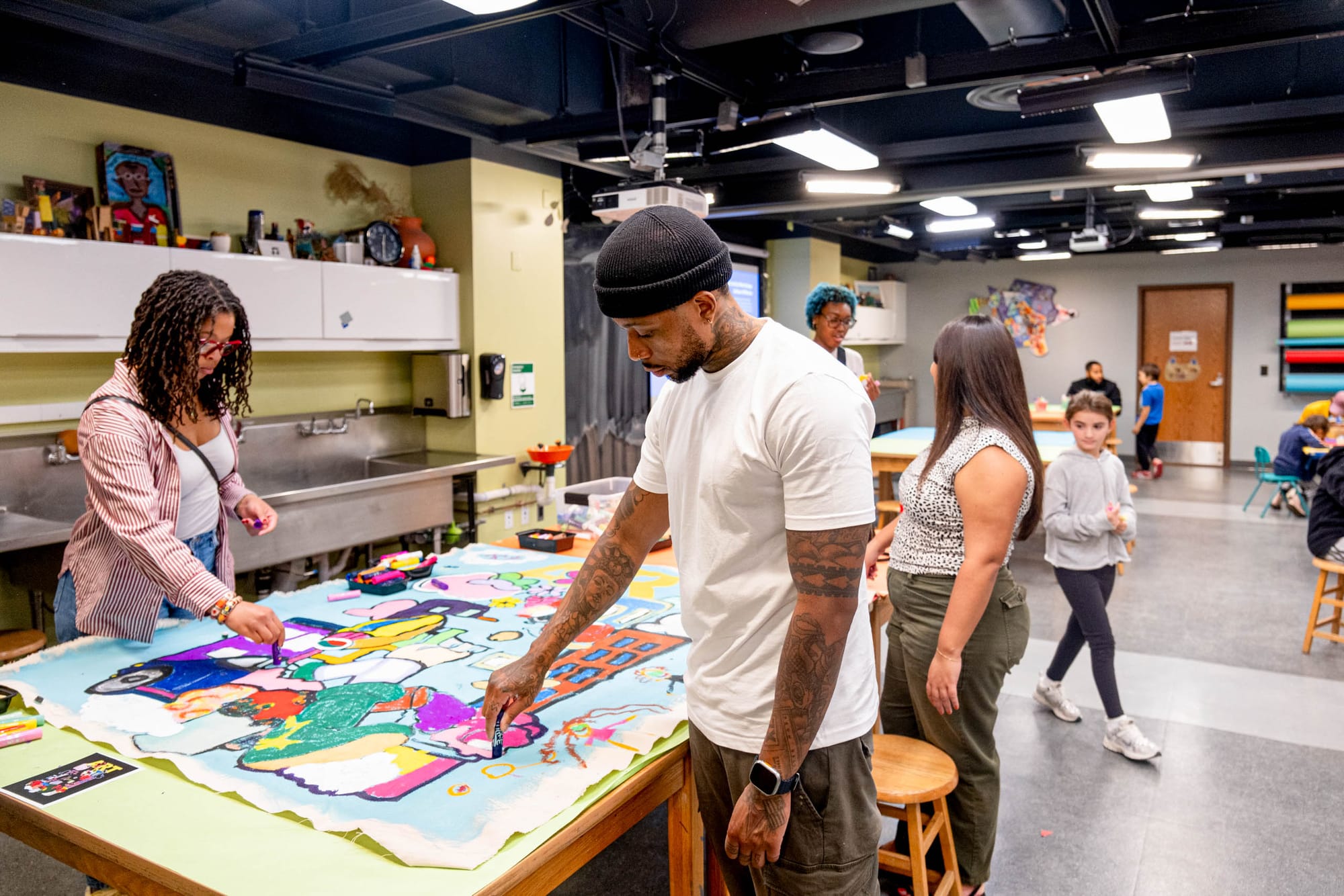 A group of adults and children work together on a colorful mural in an art classroom, using paint, brushes, and art supplies. 