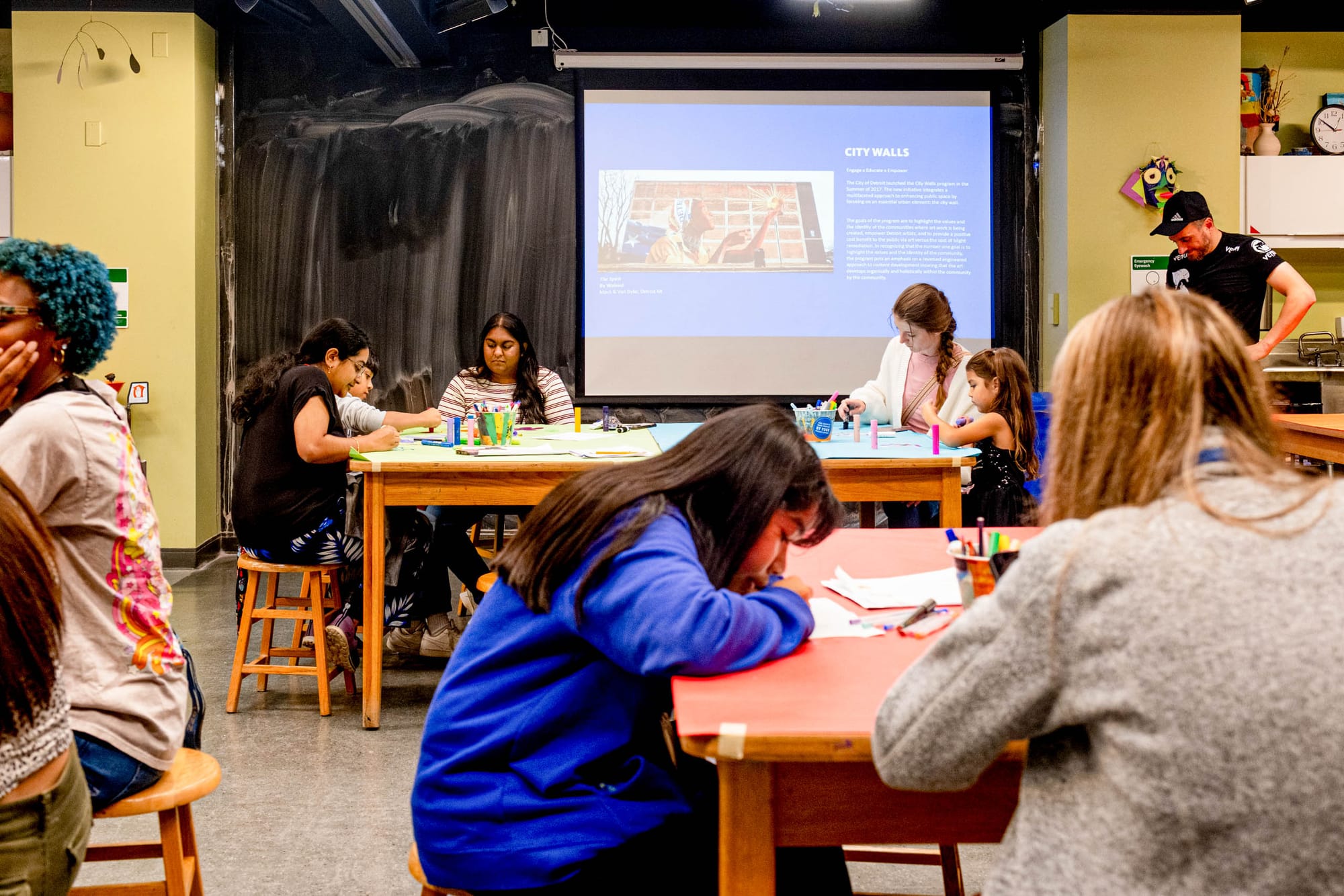 A classroom of people are drawing and sitting at tables. In the background a projector with blue slides plays. 