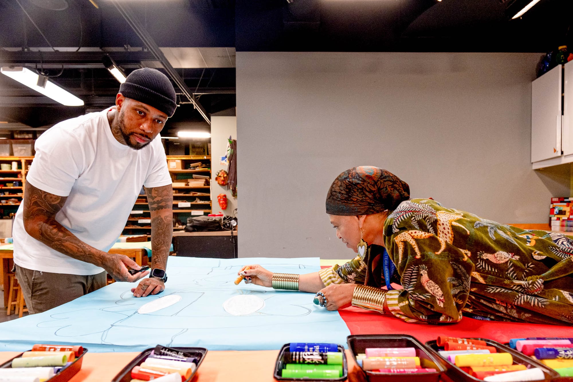 Two people, one wearing a black beanie the other wears a scarf, lean over a table collaborating on a piece of artwork. 