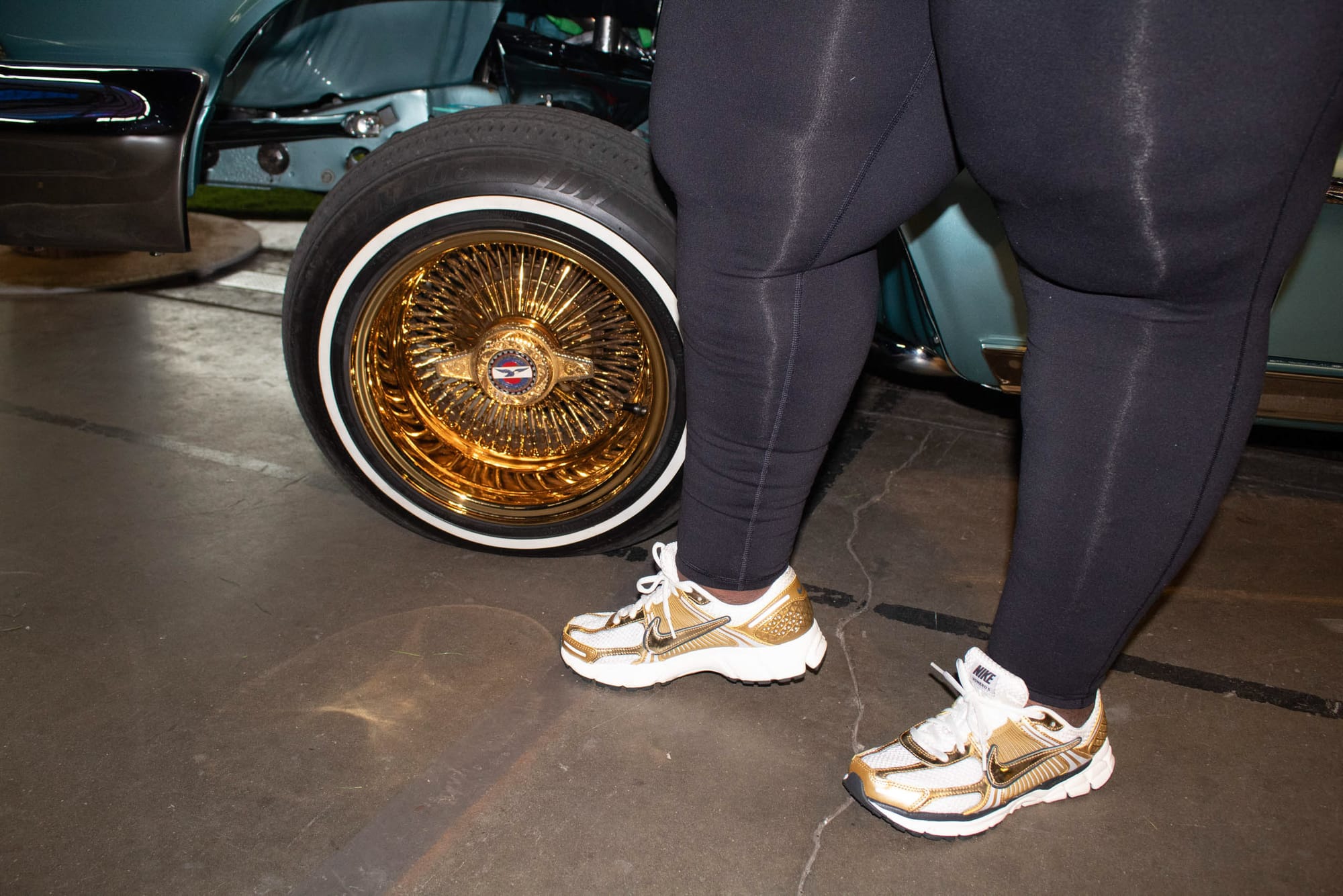 A person's lower half shows off their gold gym shoes in front of the gold rim of a classic low rider.
