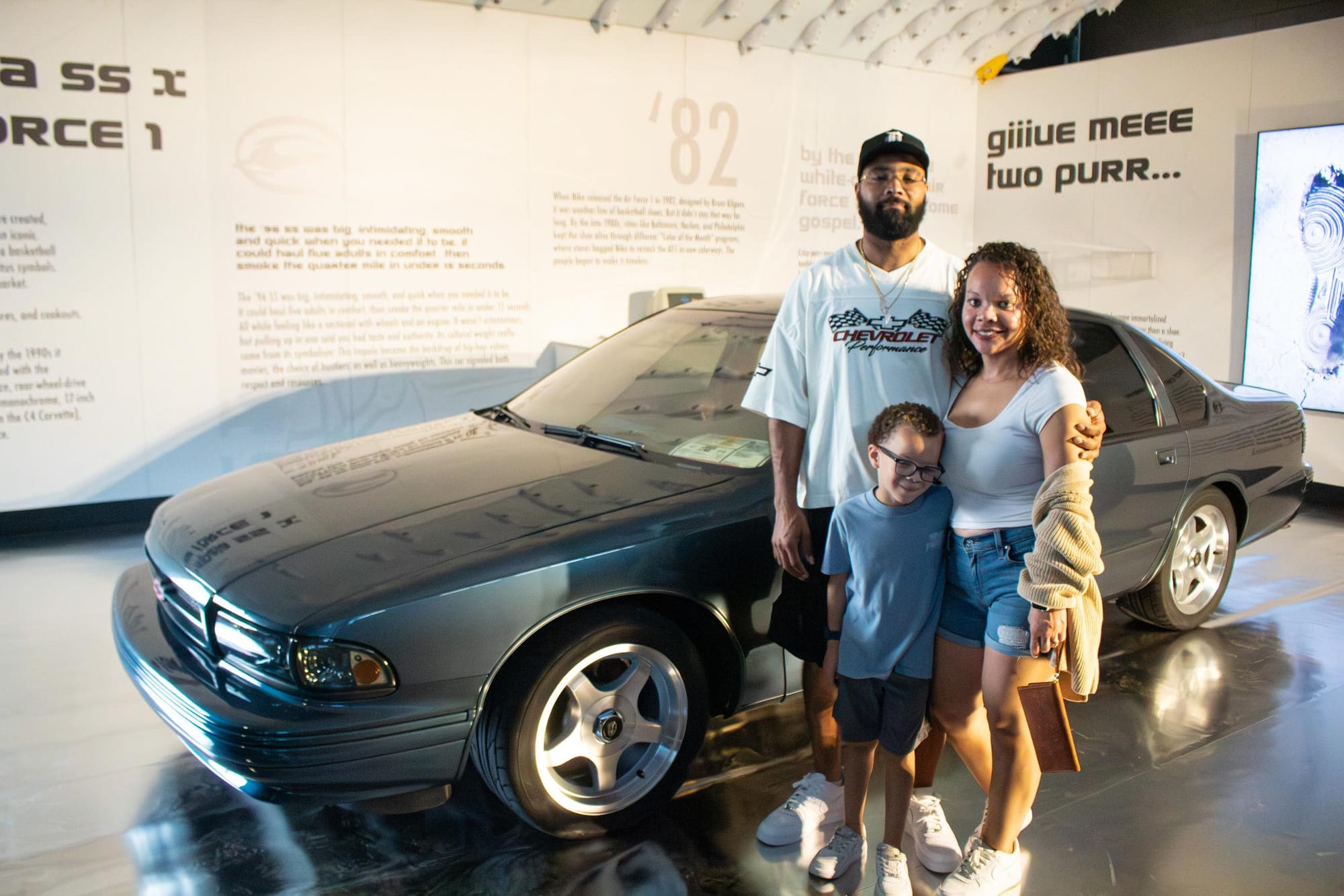 A family of three poses in front of a classic car with grey/blue paint.