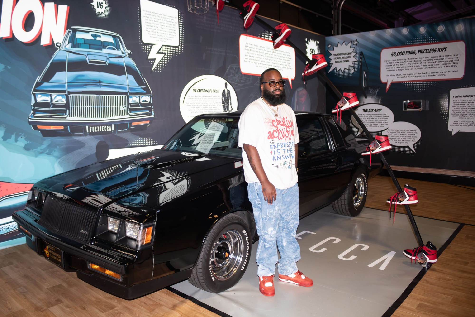 A man stands with his black classic car as basketball shoes are arranged to look like they are jumping over it.