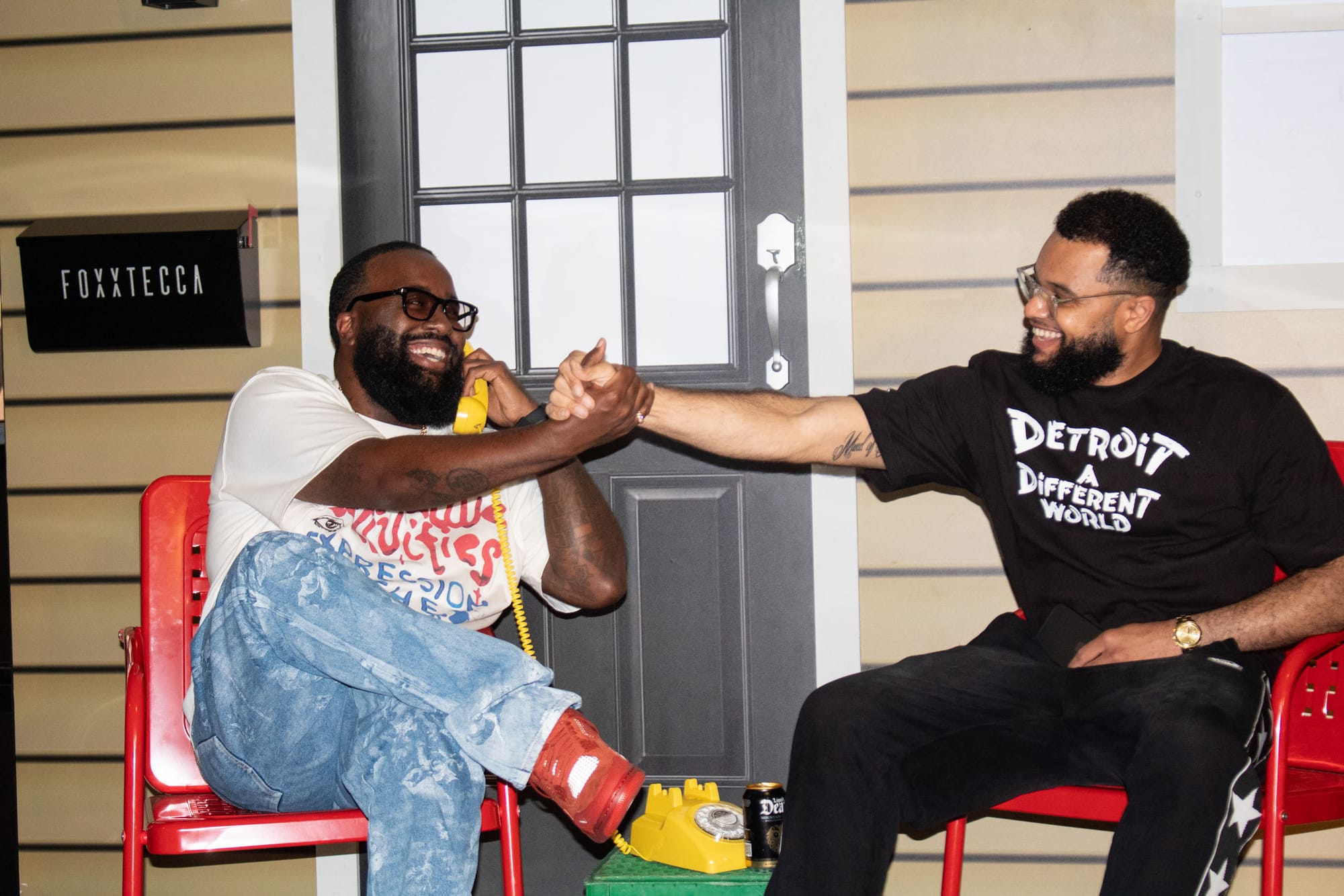 Two men sit on a staged porch, smiling giving each other a handshake. 