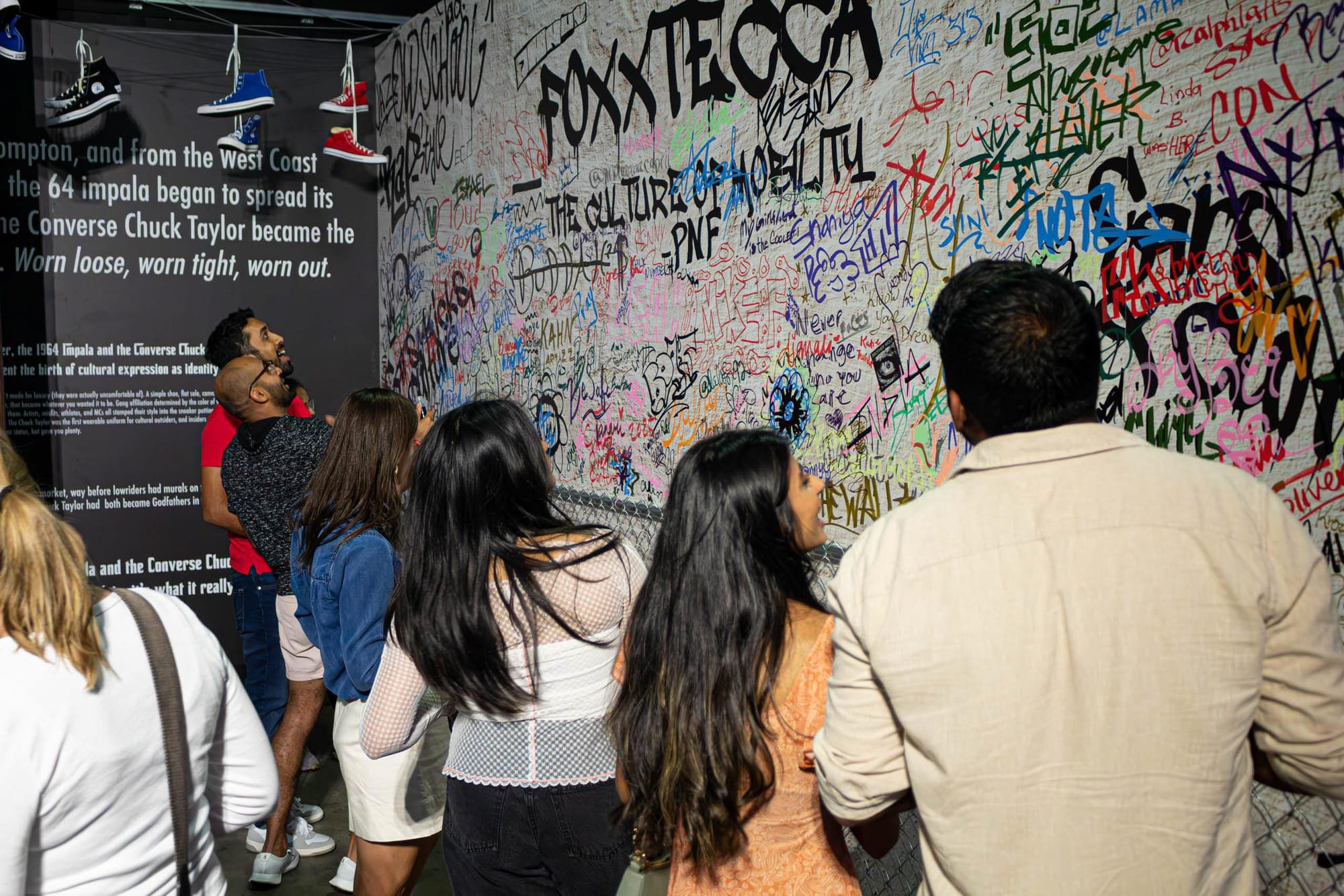 A small crowd of people gathers around a wall of graffiti-style signatures.