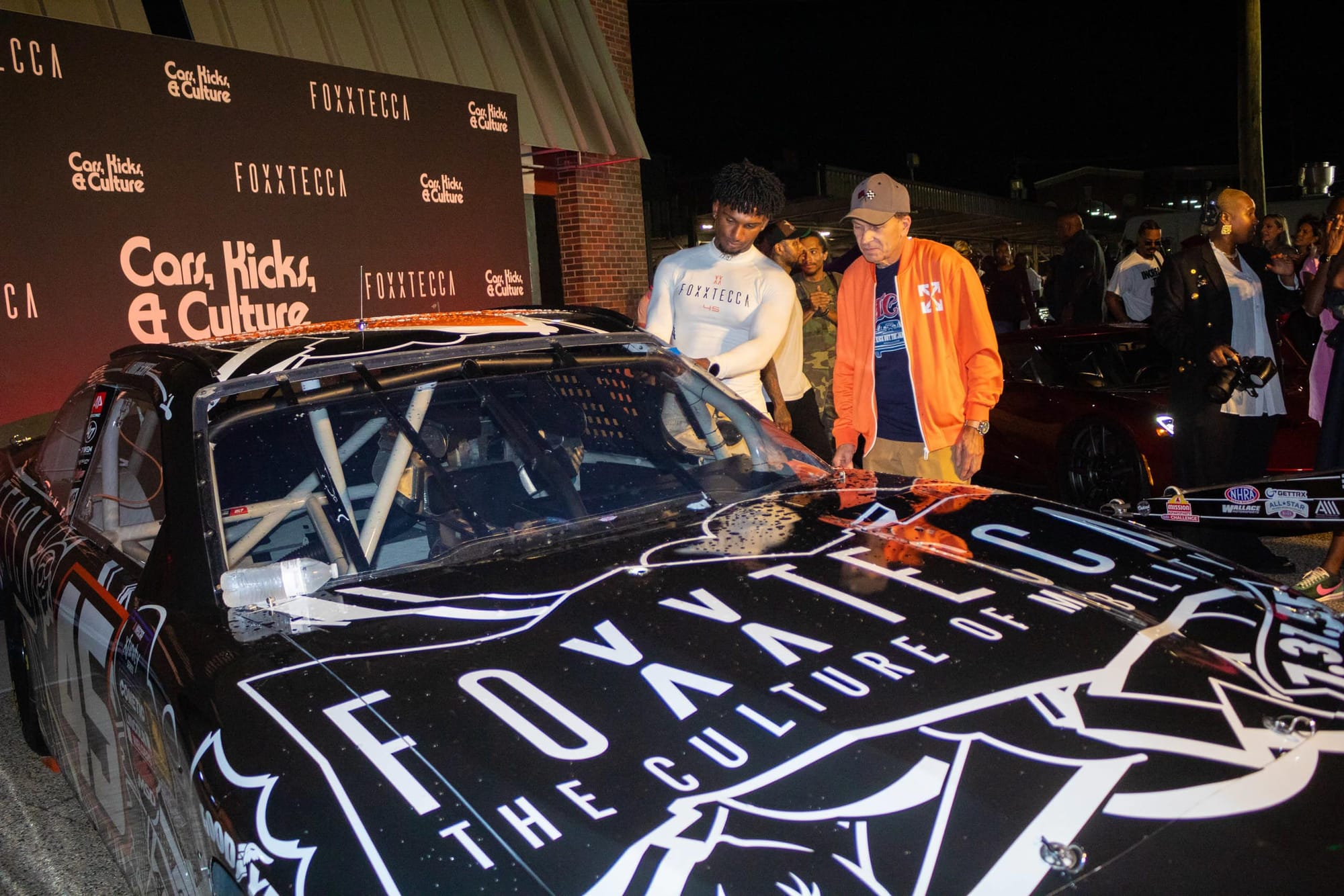 Two men examine a black race car with white lettering across the hood.