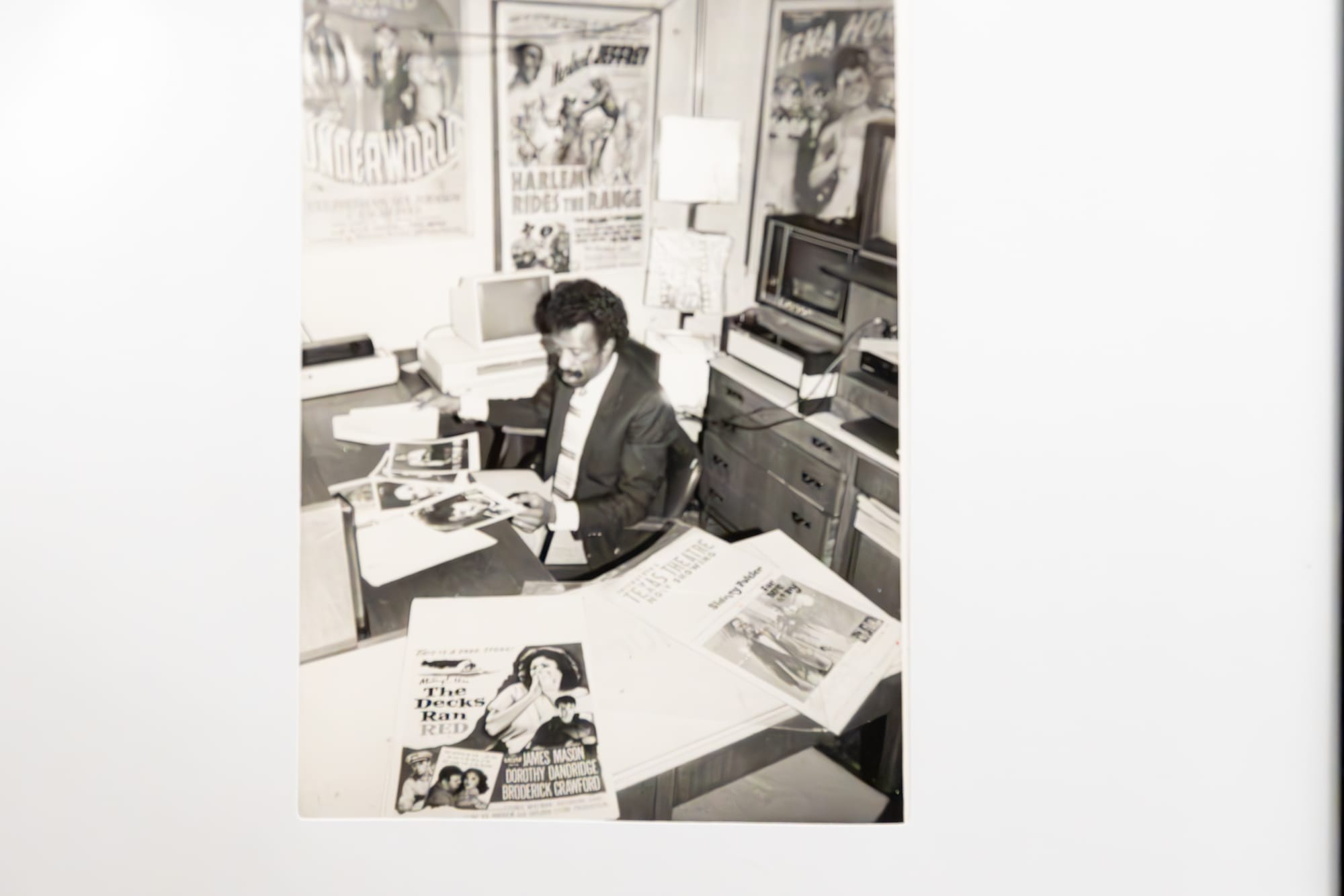 A black and white photo of man sits in his office, surrounded by posters and magazines.