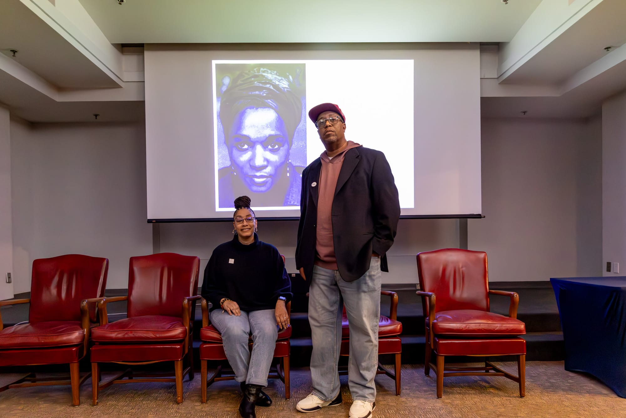 Two people stand in front of a white backdrop, a clothing rack and a poster stands on opposite sides of them. 
