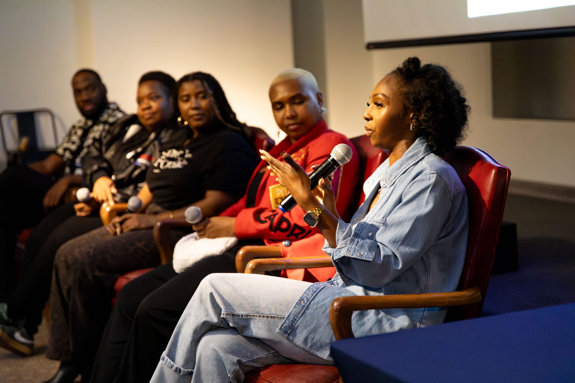 Five people sit in a row, while one person in a blue jean jacket speaks into a microphone as the others listen.