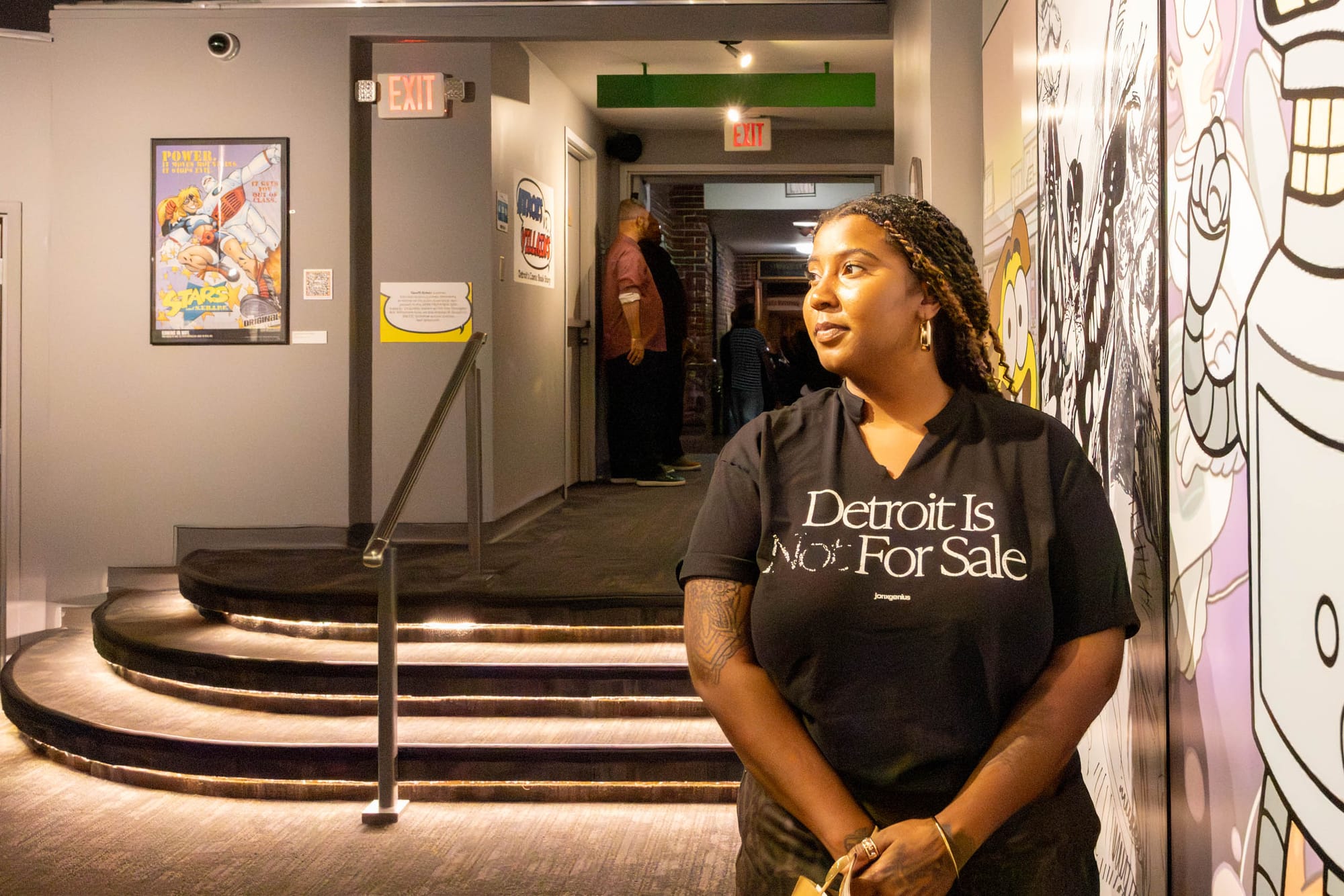 A person leans against a wall, looking to the side, wearing a shirt that reads, “Detroit Is Not For Sale.