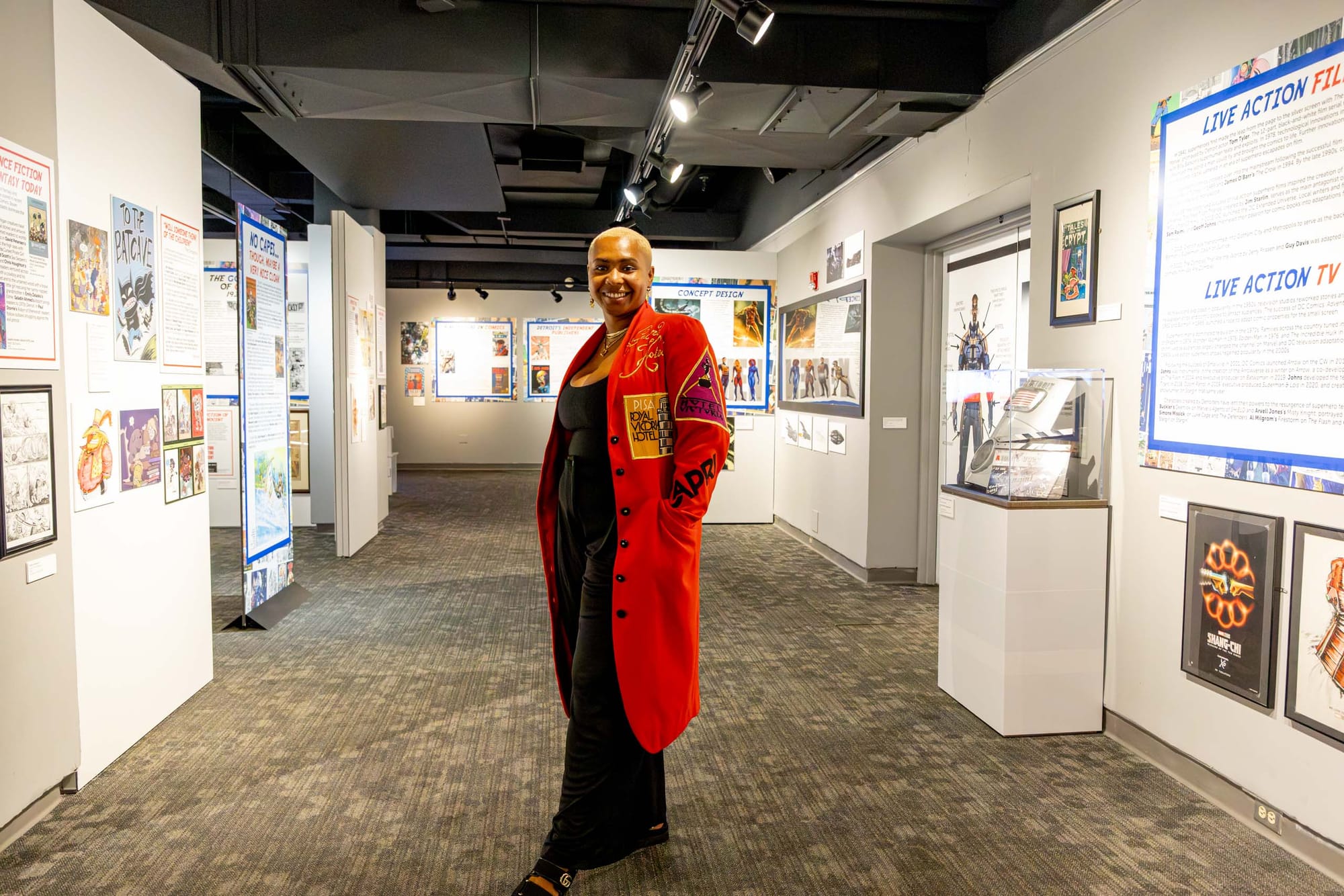 A person in a red coat stands in the center of a white room, surrounded by artworks and posters on the walls.