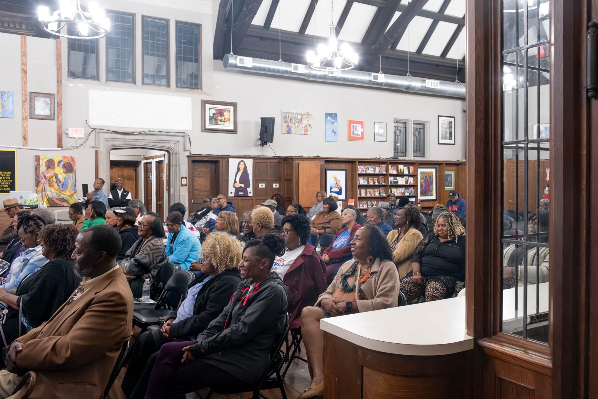 Audience members smile as they look toward the stage inside a wood-paneled room lined with bookshelves and magazines.