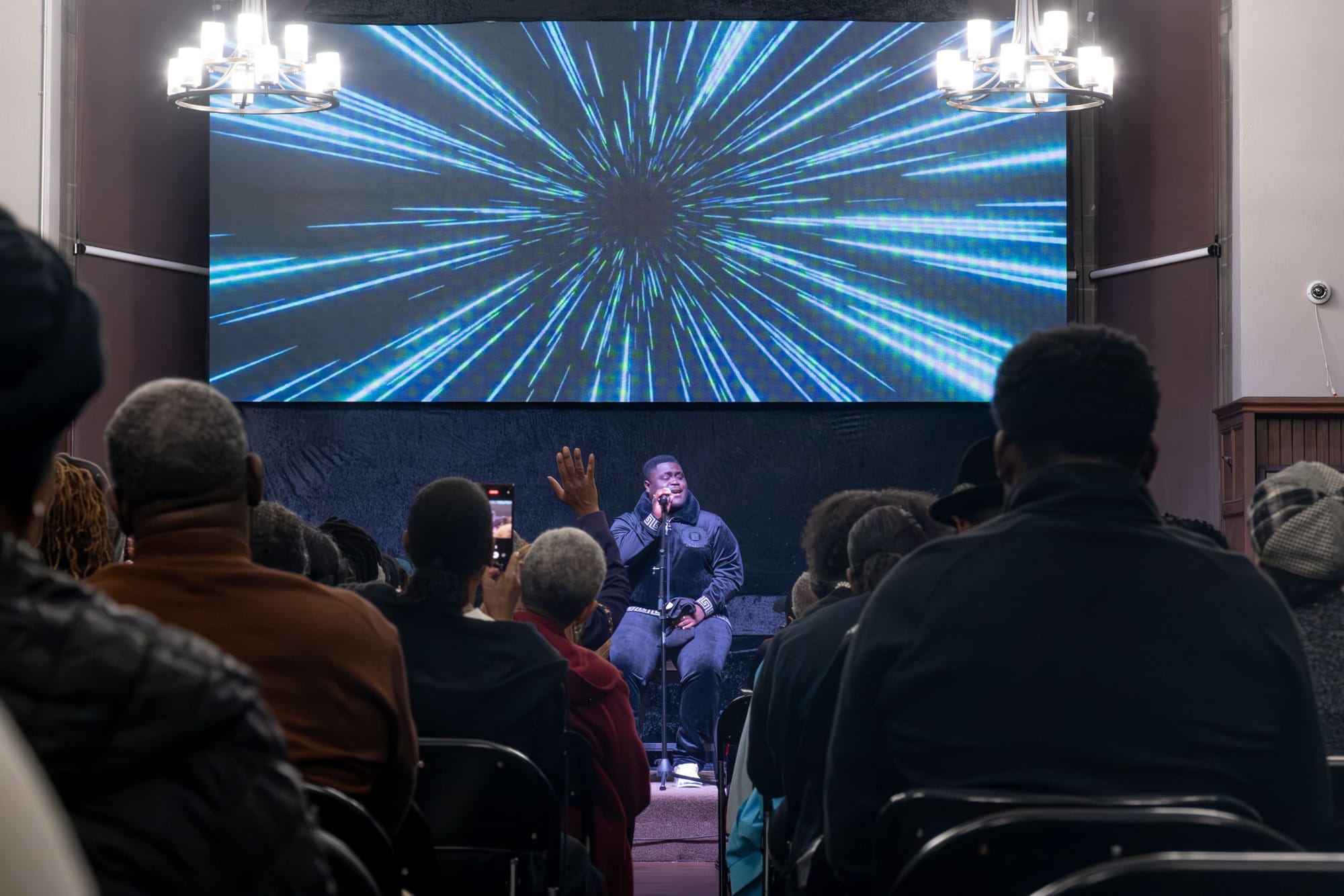 A man sits singing into a microphone, people clap in the background.