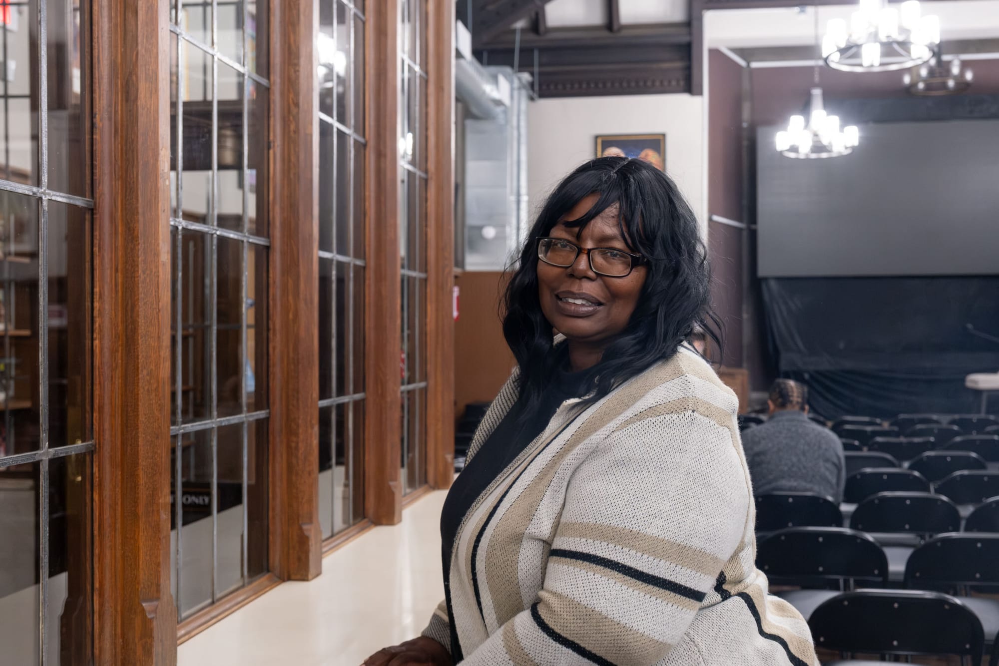 A woman in a beige cardigan and glasses leans on a table, smiling.