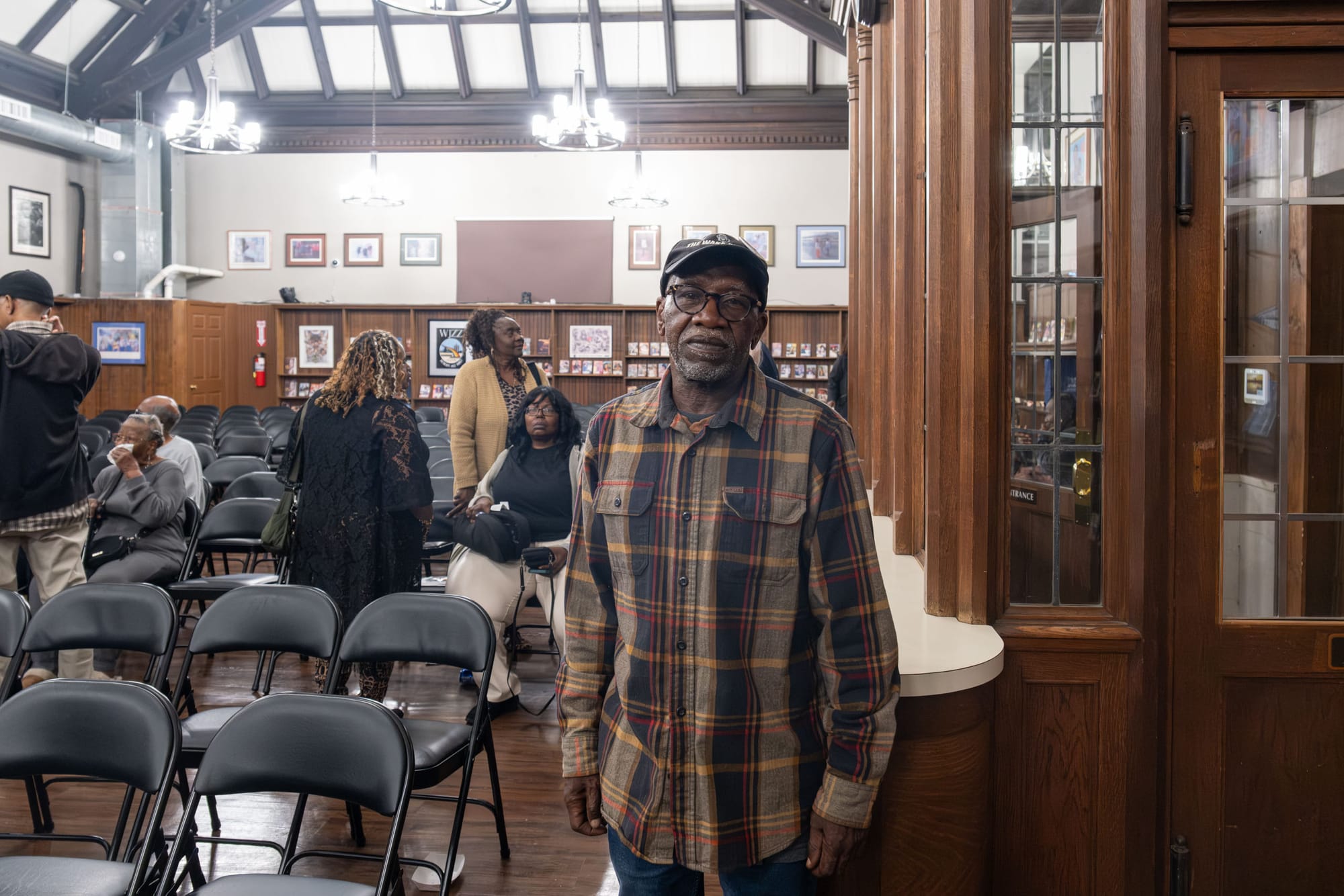 A man in a plaid shirt and glasses stands inside a room with a chandelier and wood-paneled walls.