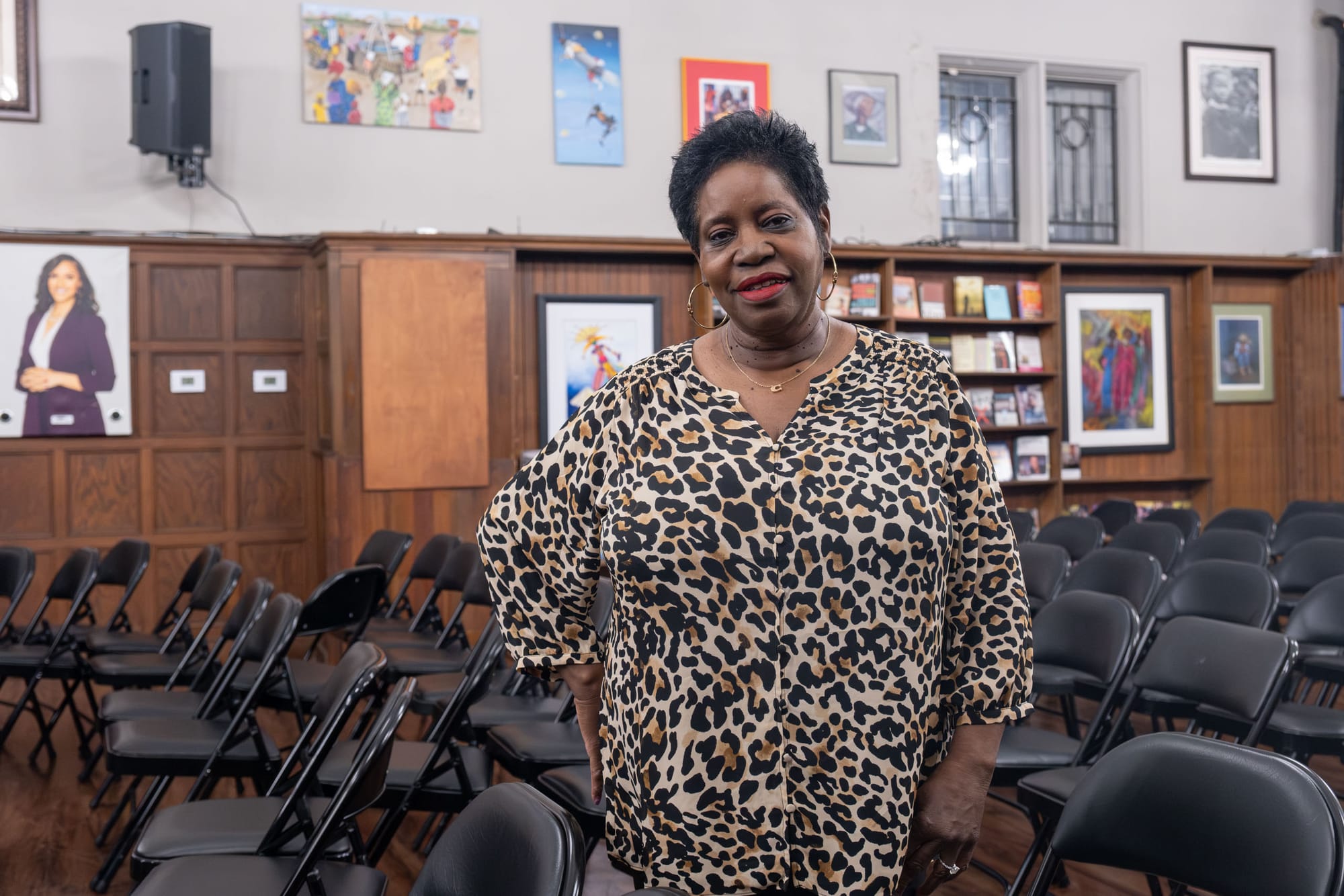 A woman in a cheetah-print shirt leans on a chair, with paintings hanging on the walls behind her.
