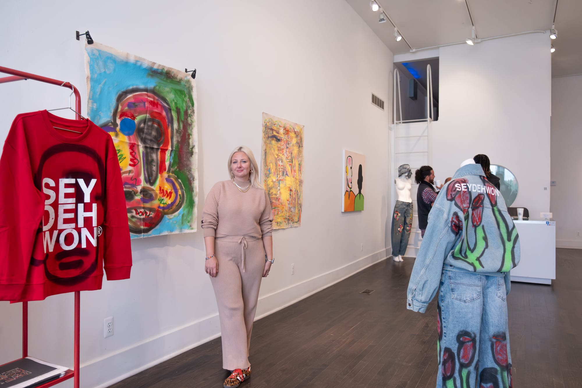 A woman wearing a tan color outfit stands in front of a collection of paintings and artworks that surround her.