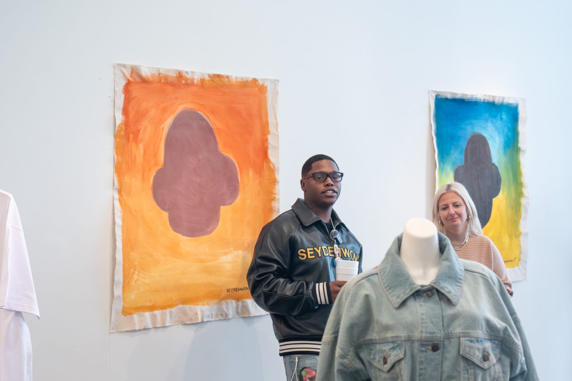 Two people stand before vibrant abstract paintings, observing a mannequin dressed in a jean jacket.