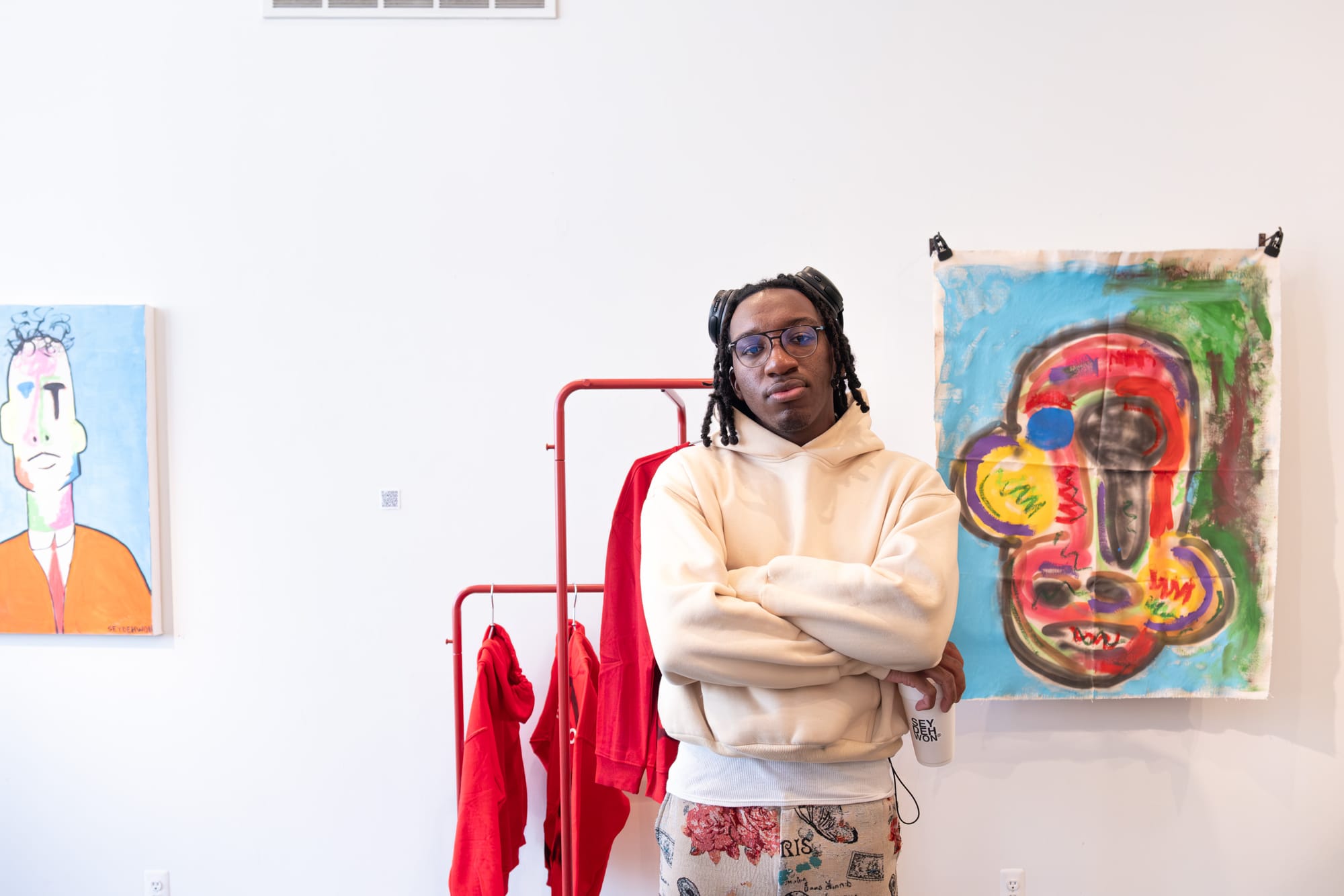 A man in a tan hoodie stands with his arms folded in front of vibrant, abstract art pieces.
