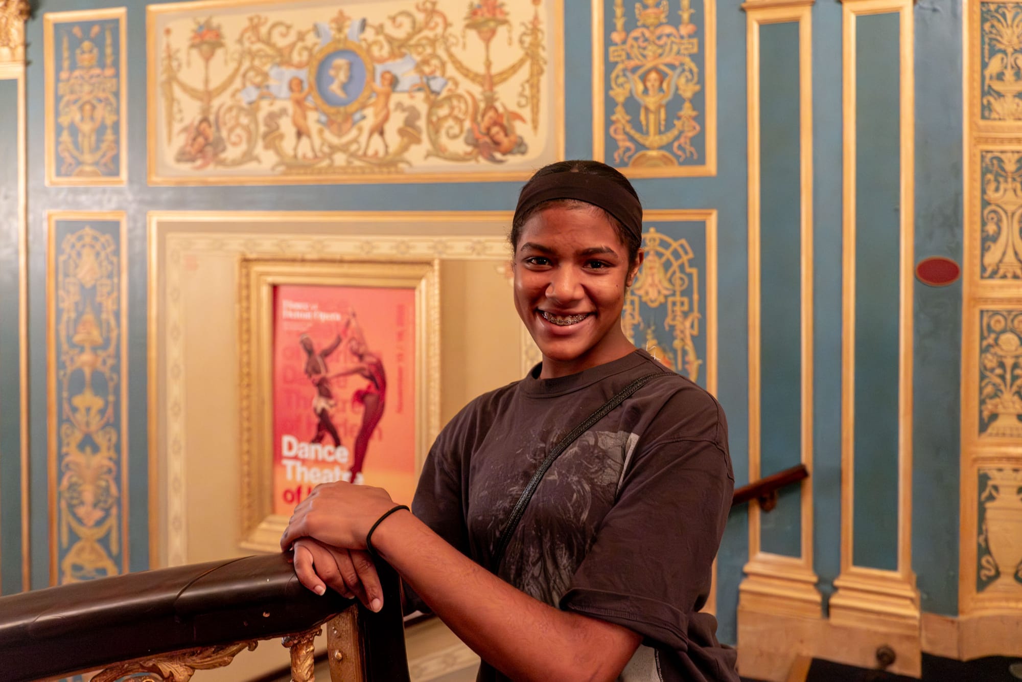 A young woman holds onto a railing in a warm room. In the background a poster reads, “Dance Theater of Harlem.”