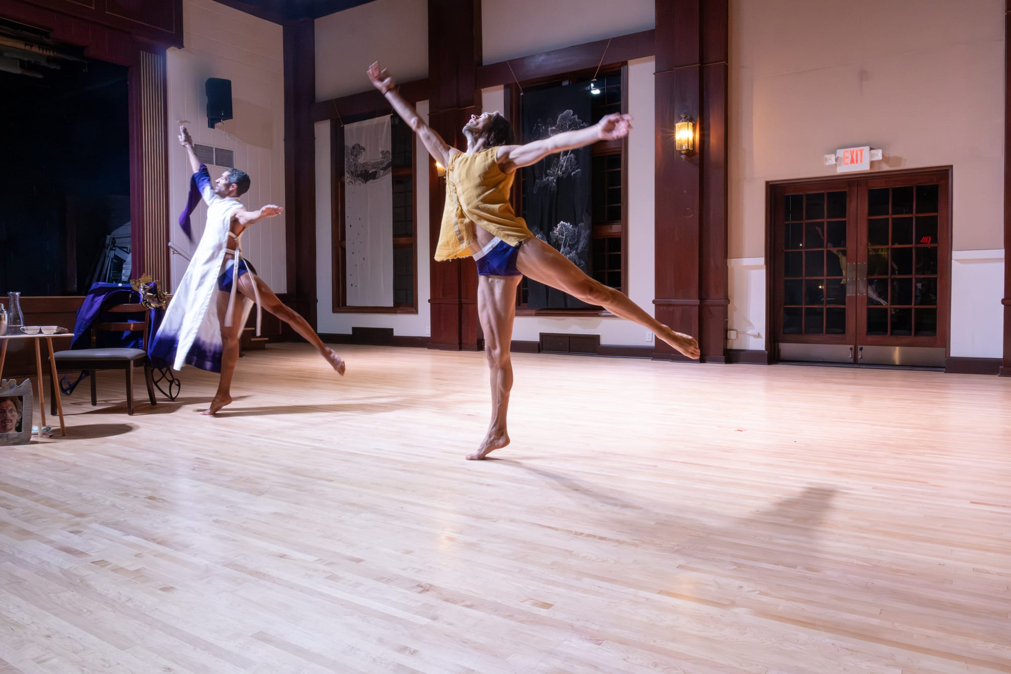 Two dancers leap into the air; one wears a purple and white gown, and the other wears a yellow top.