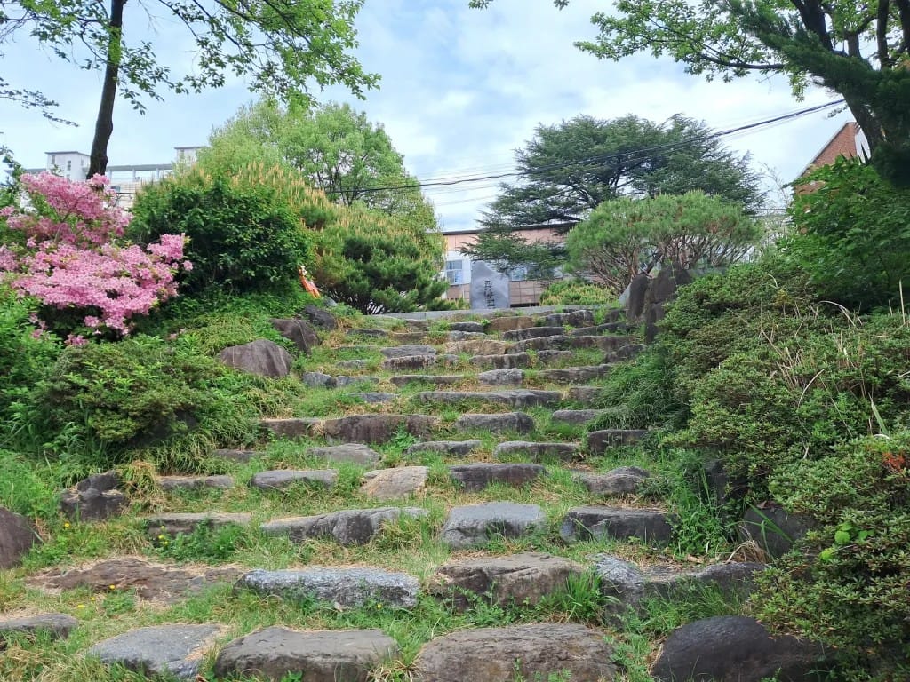 A perspective-heavy view of an outdoor scene with a lot of greenery. There are many stone steps receding into the distance. On the left, there is a pink flower bush. Way in the back, you can see a large stone monument.