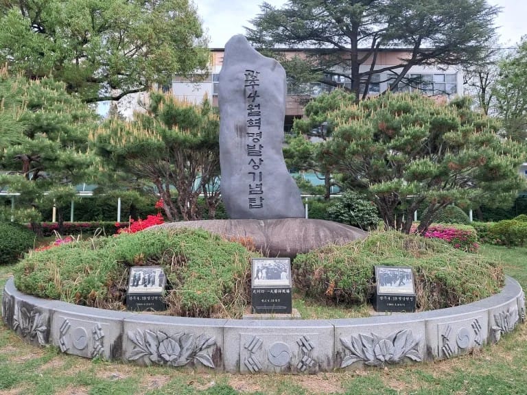 A large rectangular stone, standing vertically. There are Korean letters inscribed on it. In front are three photographs on stone markers. The whole scene has a lot of bushes and trees.