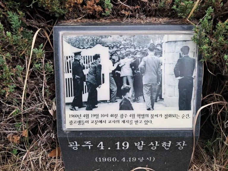 A black and white photograph affixed to a stone, nestled among greenery. In the photo, police stand by a large gate and a man in a suit throws his body against a crowd of students in school uniforms.