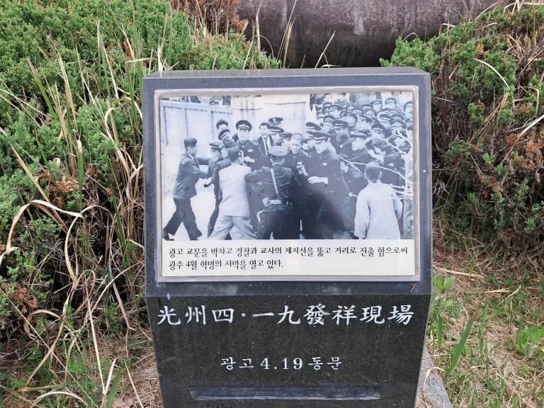 A black and white photograph affixed to a stone, nestled among greenery. In the photo, the students are breaking past the barrier of cops and teachers and are spilling out onto the street.