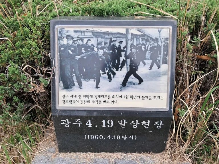 A black and white photograph affixed to a stone, nestled among greenery. In the photo, some people look confused, one young man is running, focused, and a couple cops are following behind.