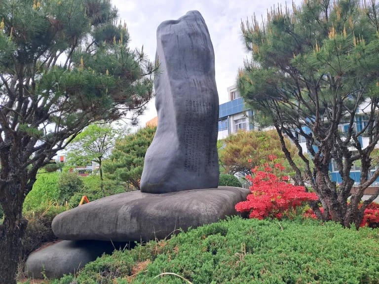 A "back" view of the large rectangular stone monument, which shows writing in smaller type than the front. There are pine trees on either side and more bushes and flowers. From here you can see the stone is resting on another, horizontal stone.