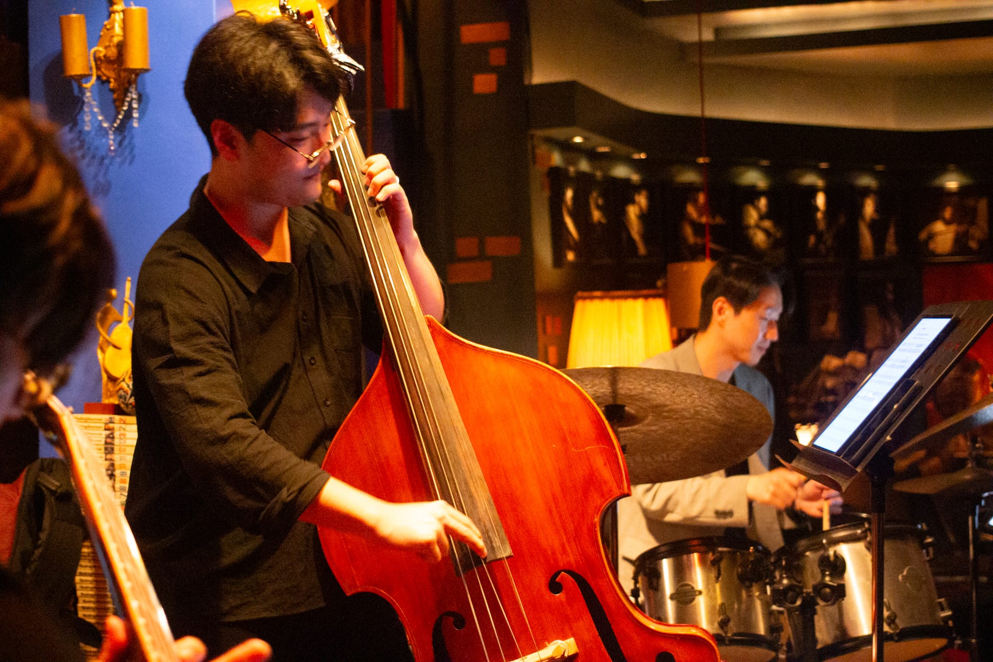 Interior of a music venue with a bassist and drummer. In the distance is a section lined with framed photos of jazz musicians. The lighting is warm and dim.