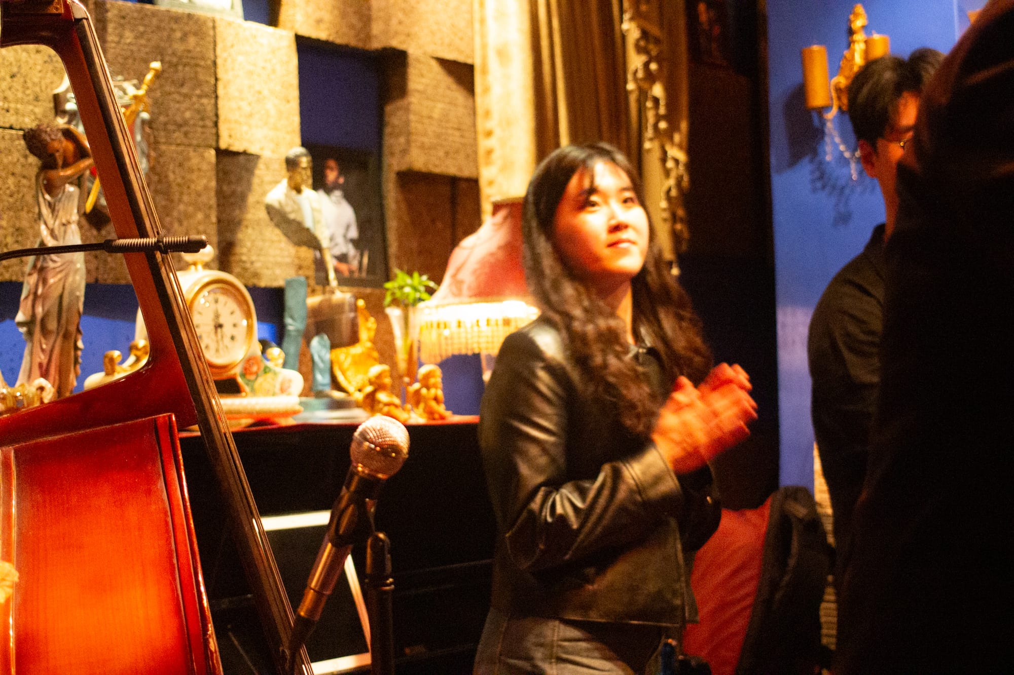 Interior of an eclectic music venue. There is a clock and some figurines on the piano. Joo Jeong stands up, clapping.