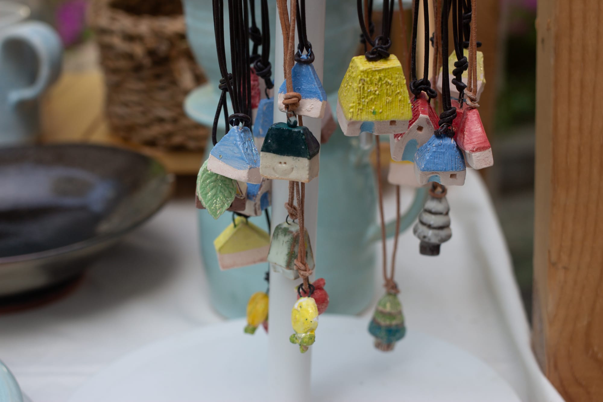Several small ceramic charms on necklaces, on display. Most are little houses, some with faces. They are in cheerful colors, yellow, green, pink, blue, and off-white.