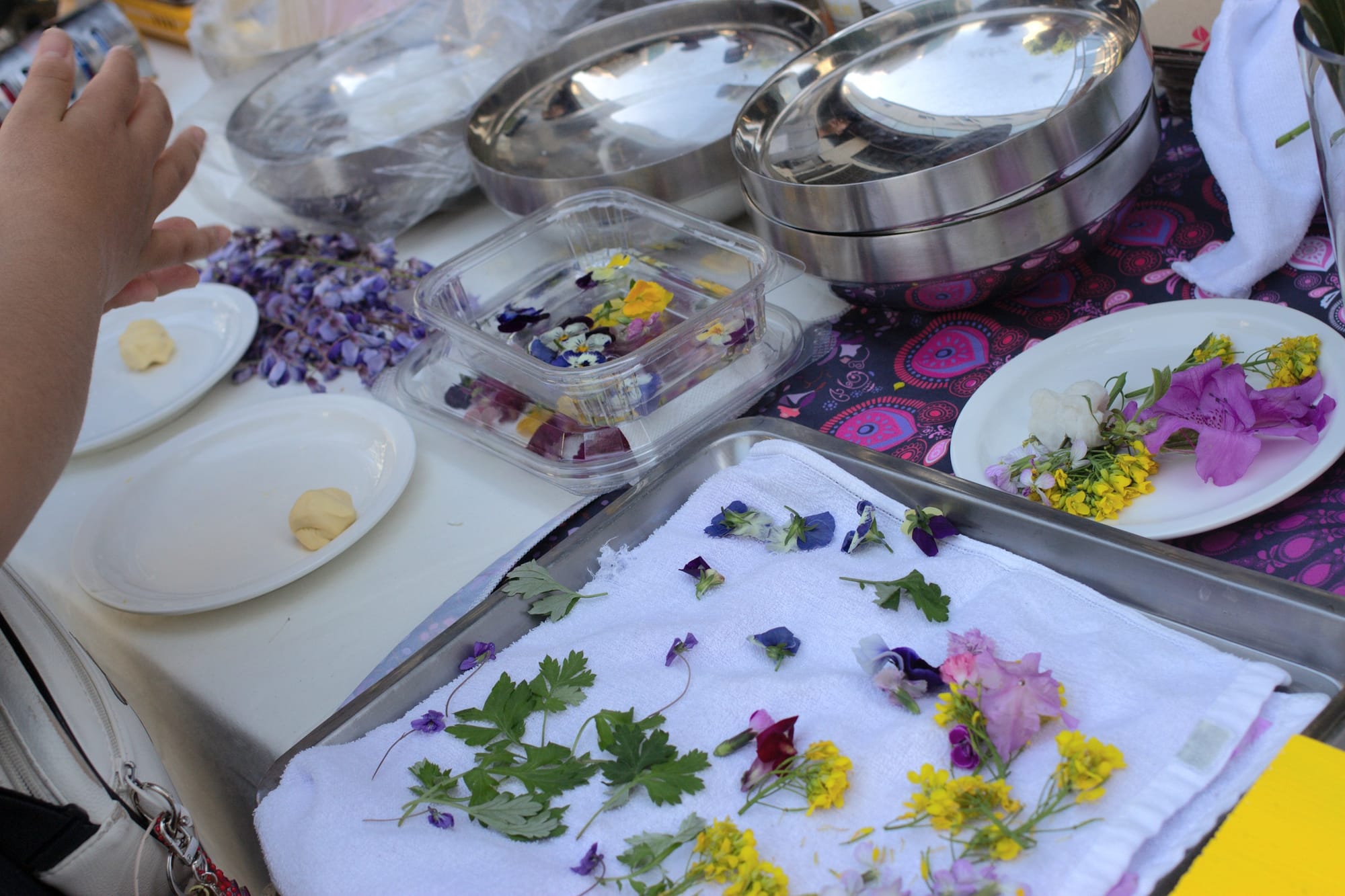 Several small flowers are drying on a white towel. A hand is forming some dough. There are some dishes and metal bowls on the table.