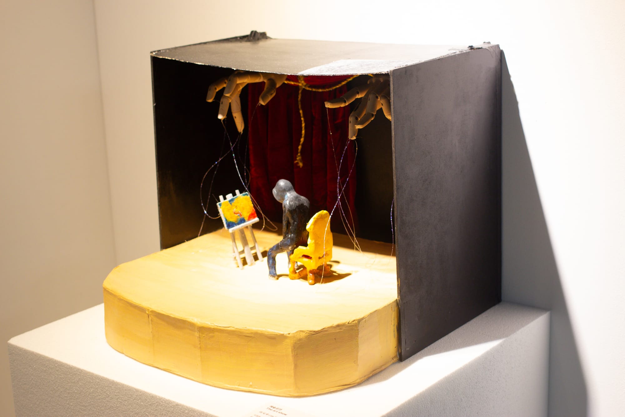 A diorama sculpture with omious hands pulling the strings. A hooded figure on stage sits in front of a painting.