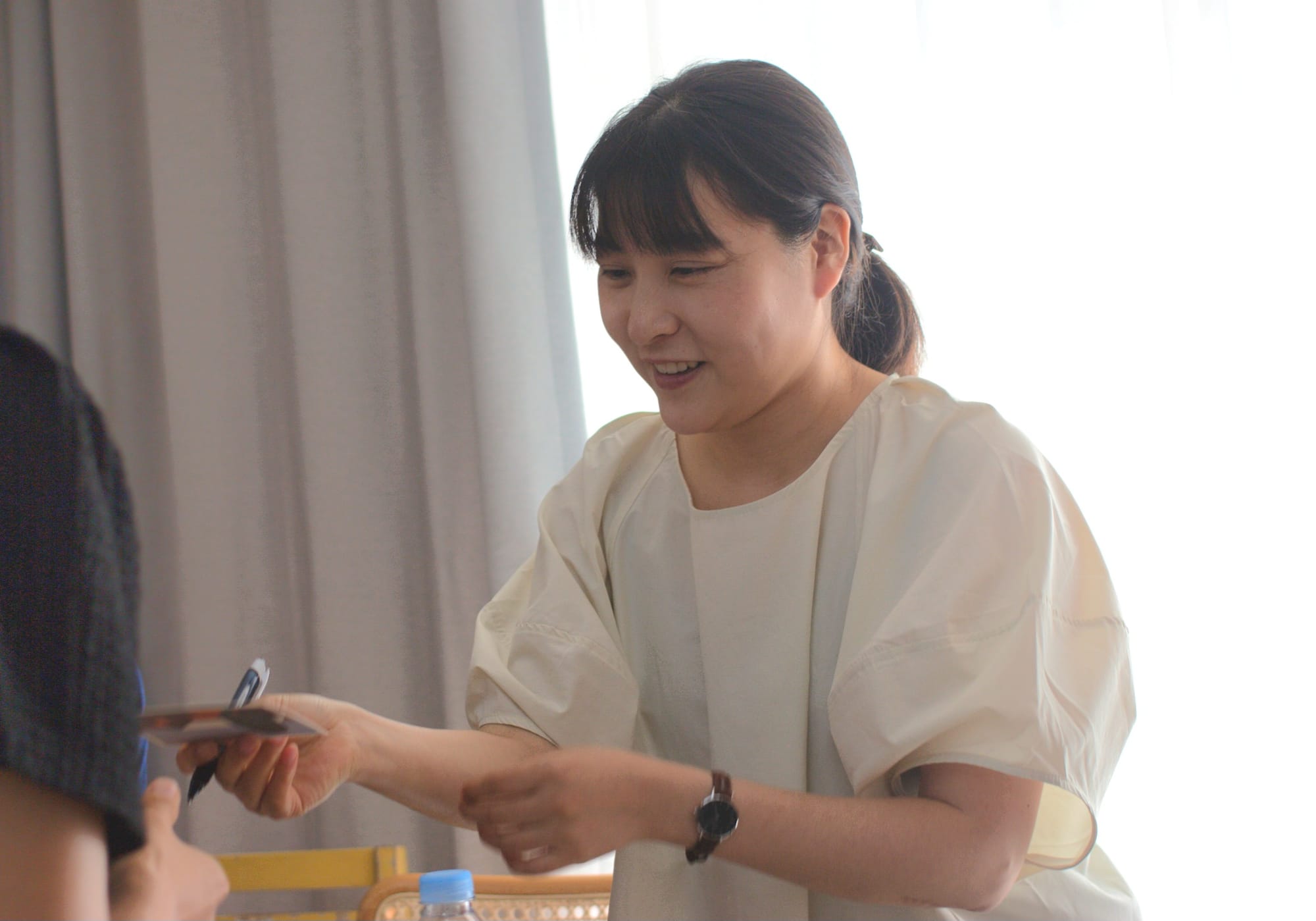 A smilong woman holds a pen and hands a signed postcard to someone. Behind her the sun is shining brightly through a window.