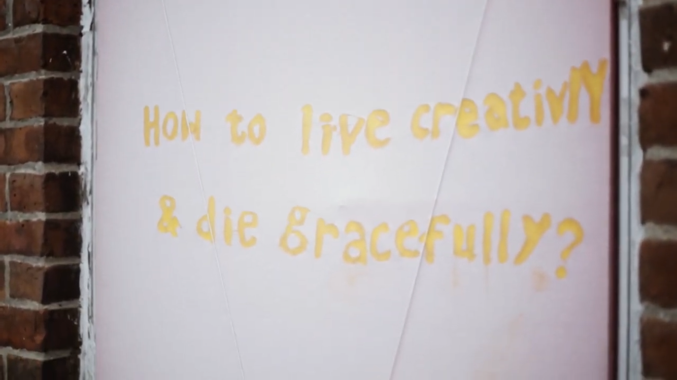 Yellow words painted on a lilac background on a building exterior, saying, "How to live creativly & die gracefully?"