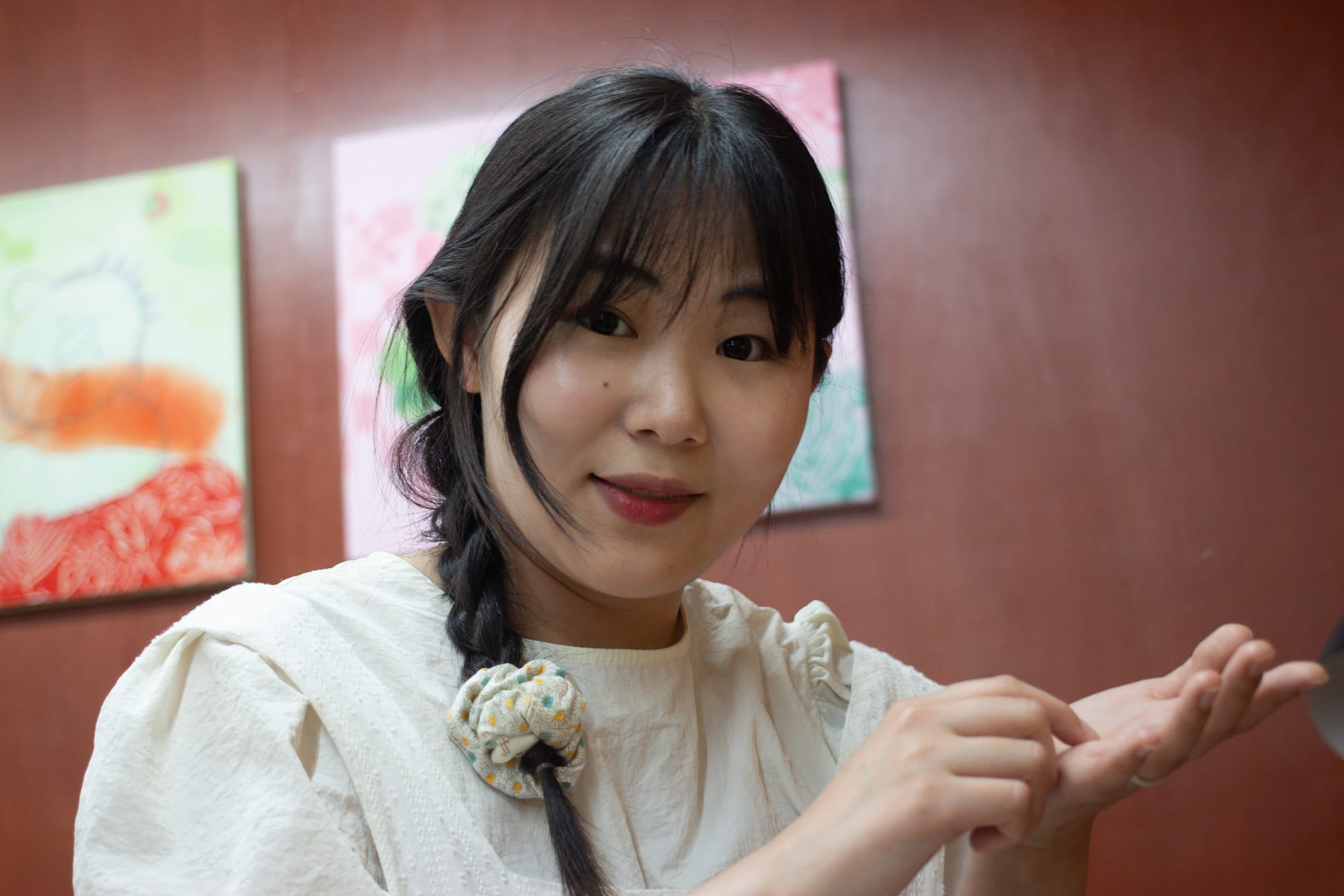 Seo Eun Ji gestures with her hands. One hand is pinched, and the other is out flat, palm up. Seo's hair is in a braid. Behind her are some paintings in pastel colors.