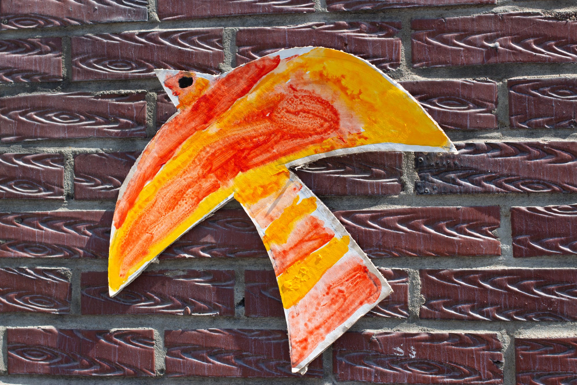 A yellow, orange, and red cut-out drawing of a bird is on an exterior brick wall. The bird has simple shapes and a single dot for an eye.