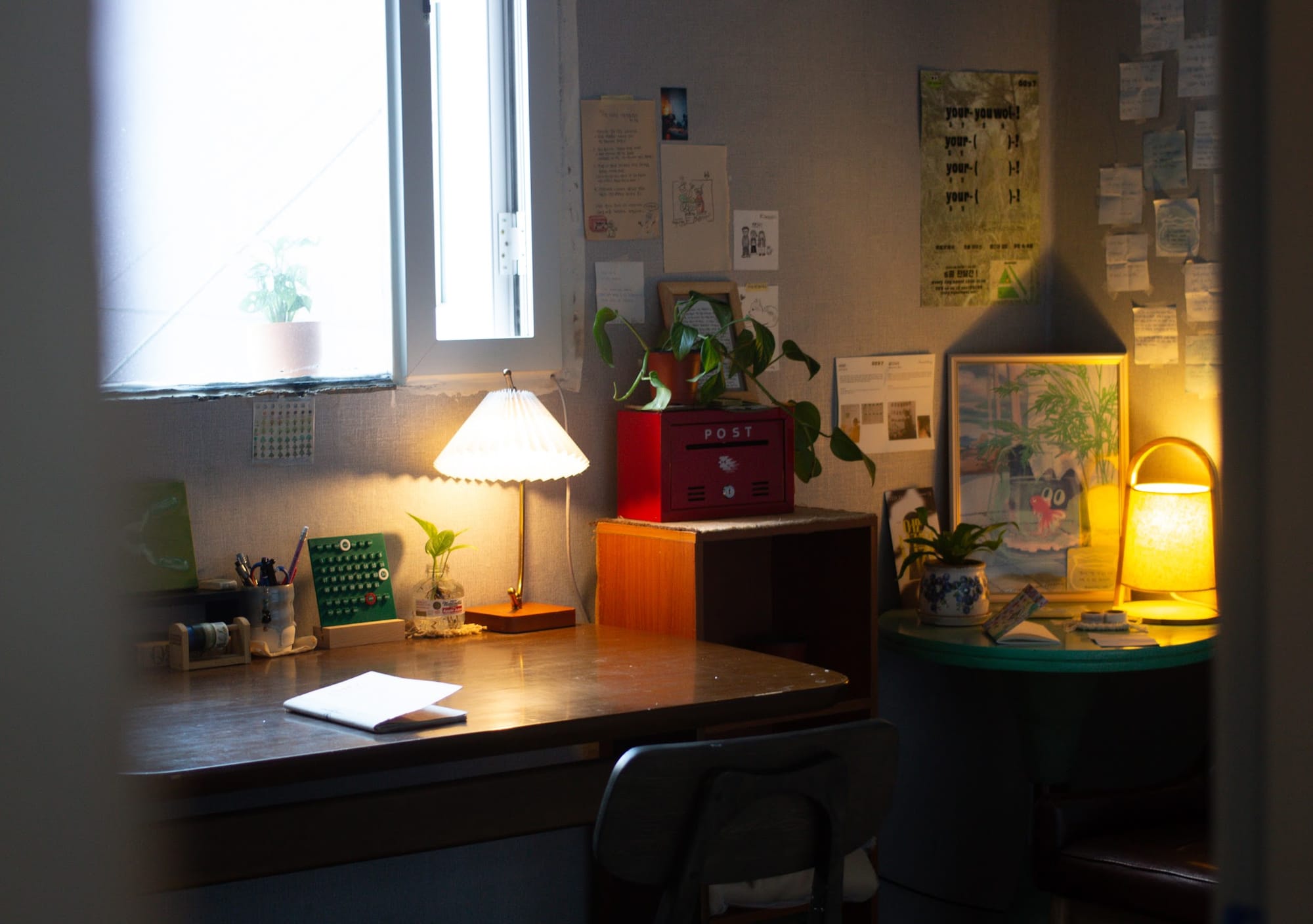 A picture taken from a doorway, of a dimly lit ccozy room with a desk, chair, lamps, plants, and a red metal postal box.
