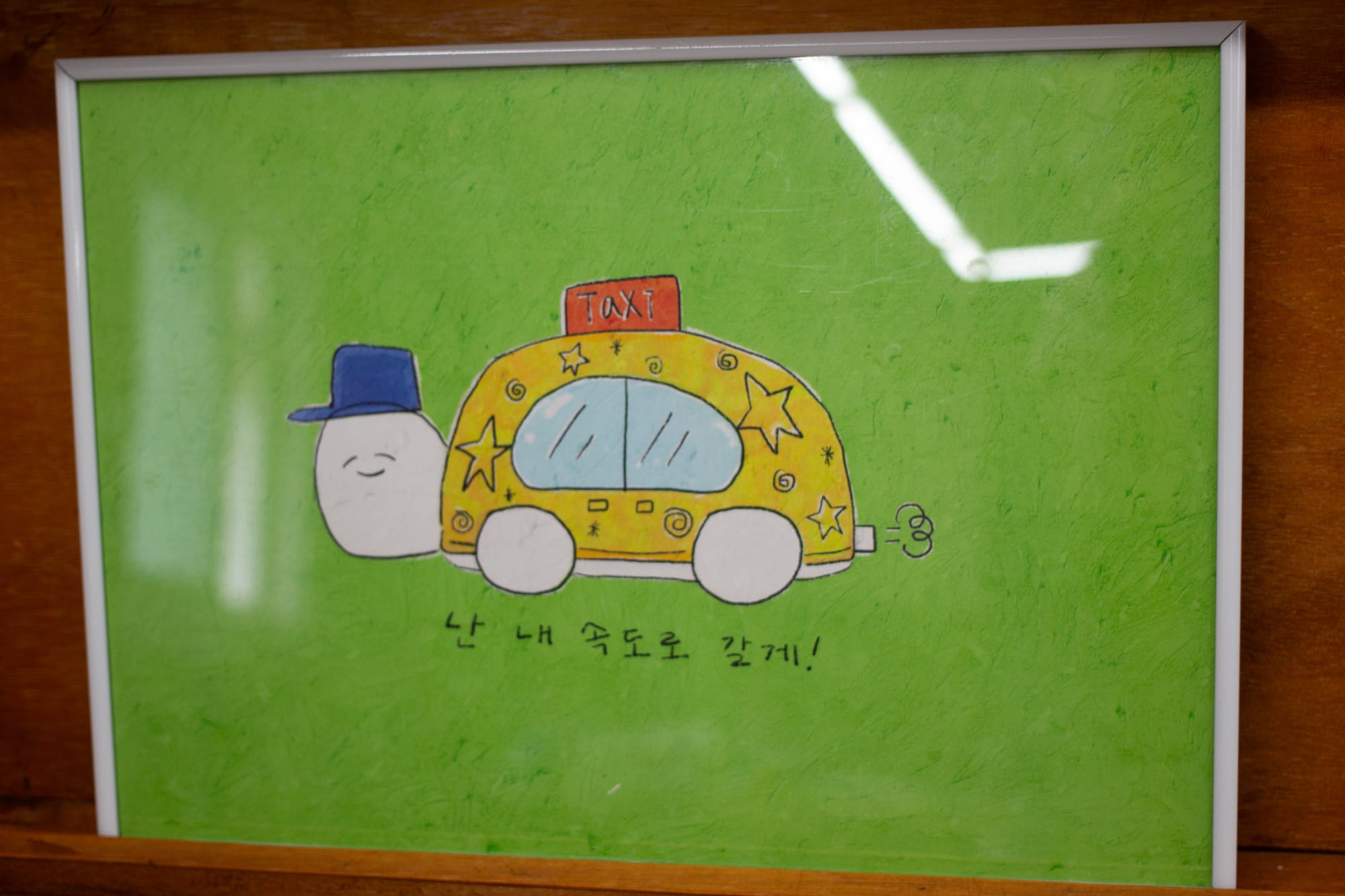 A framed drawing of a turtle that is also a taxi. It has a face, a hat, and a suitcase strapped to the top. There is a phrase in Korean: 난 내 속도로 갈게!