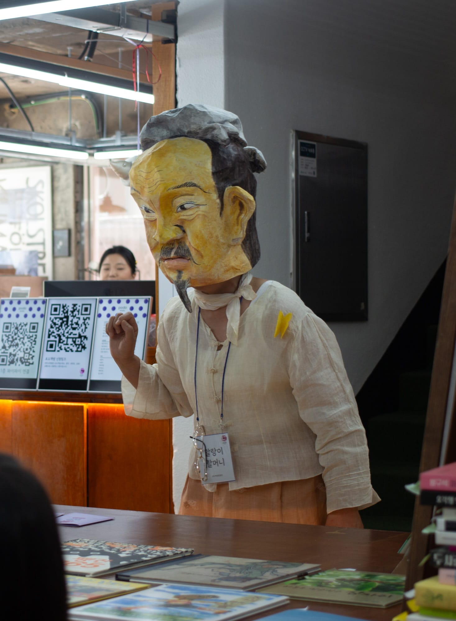 A woman in a big papier-mache mask (depicting the face of an old man) and linen clothing addresses the crowd.