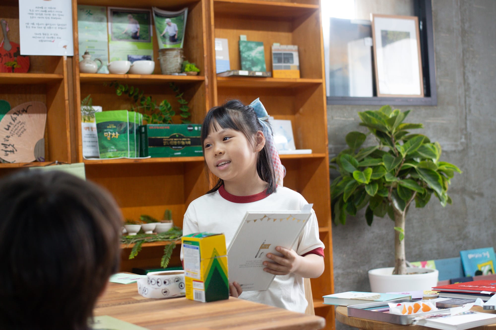 A girl with a big ribbon in her hair holds a picture book inside a bookstore. On the table in front of her are crayons and a roll of stickers with eyes.