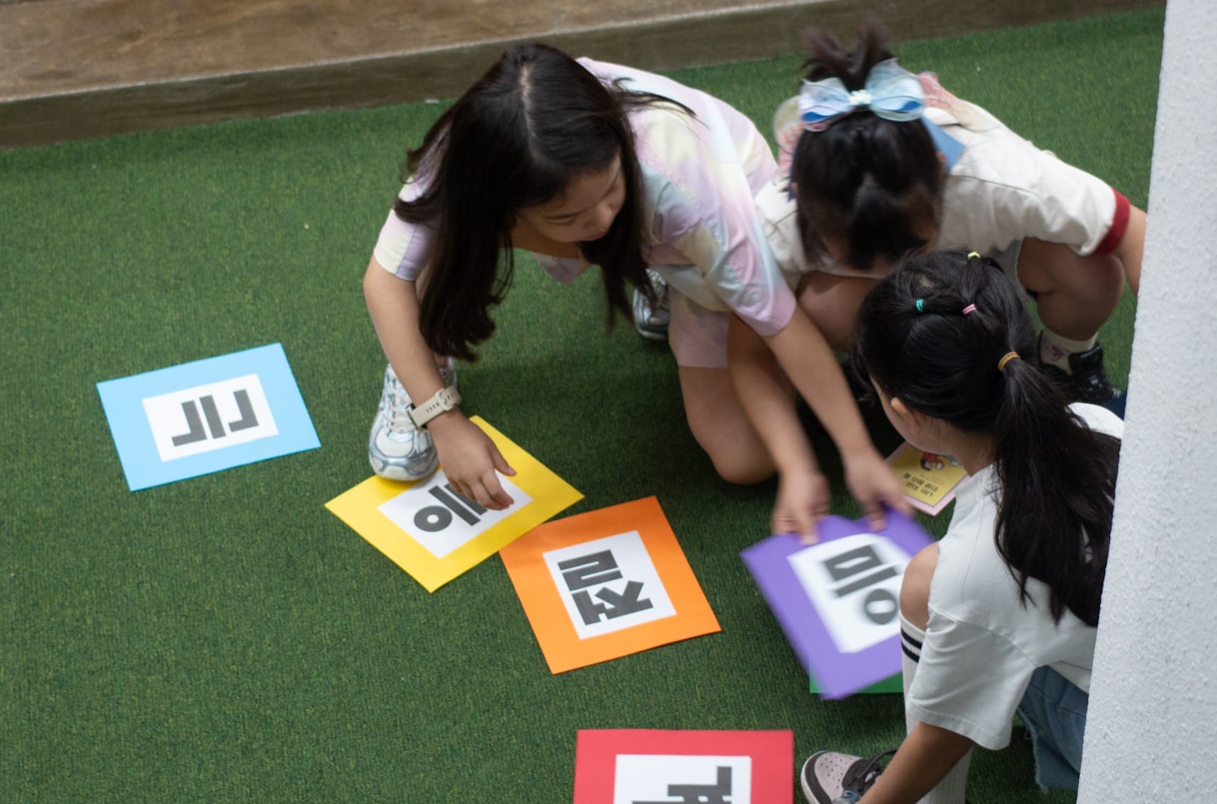 Three girls crouch on a grass-like floor, concentrating on pieces of paper with different colors and letters. They are rearranging the papers.