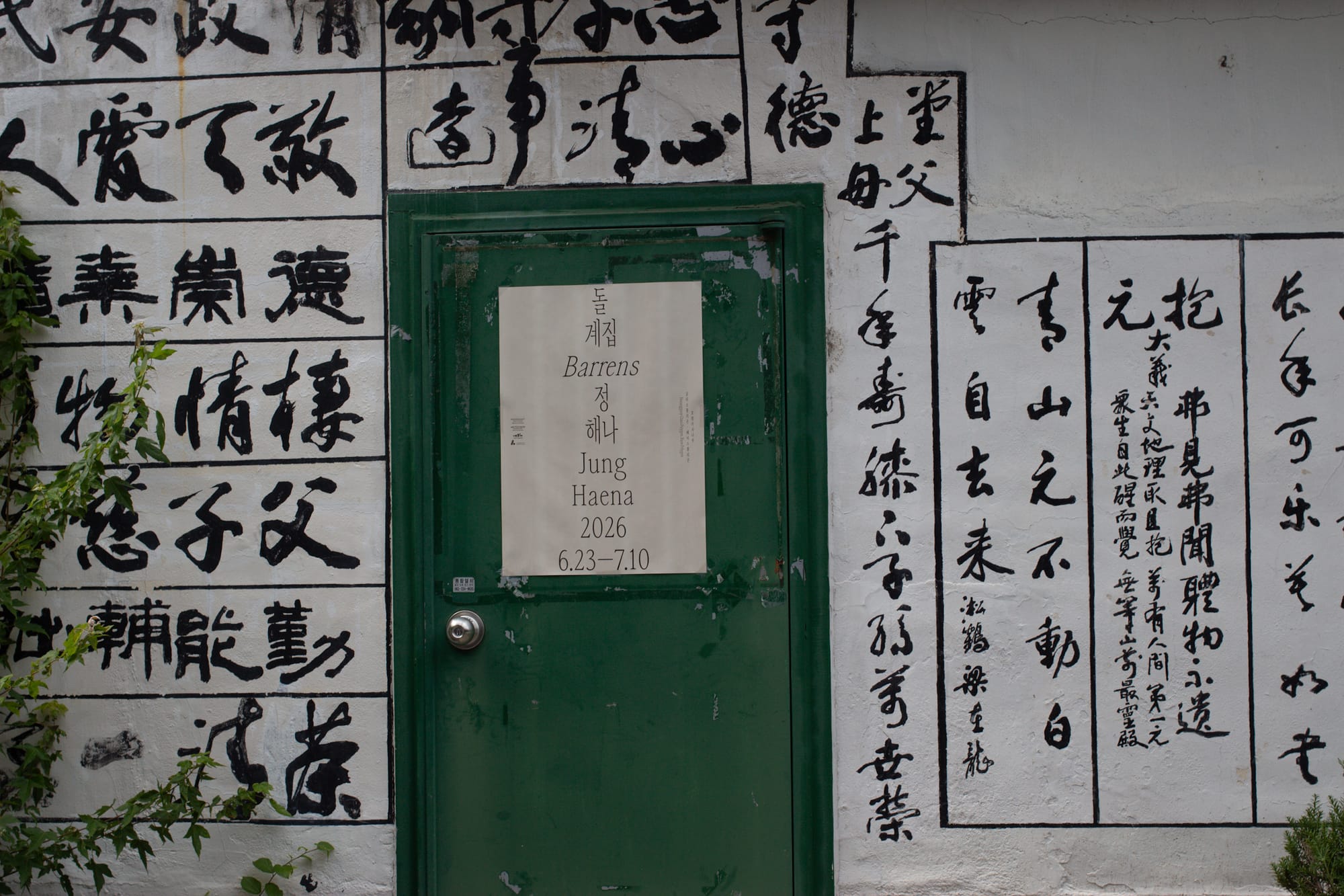 Exterior white-painted wall with calligraphy in black ink. On a green door is a large flyer for the art show Barrens by Jung Haena.