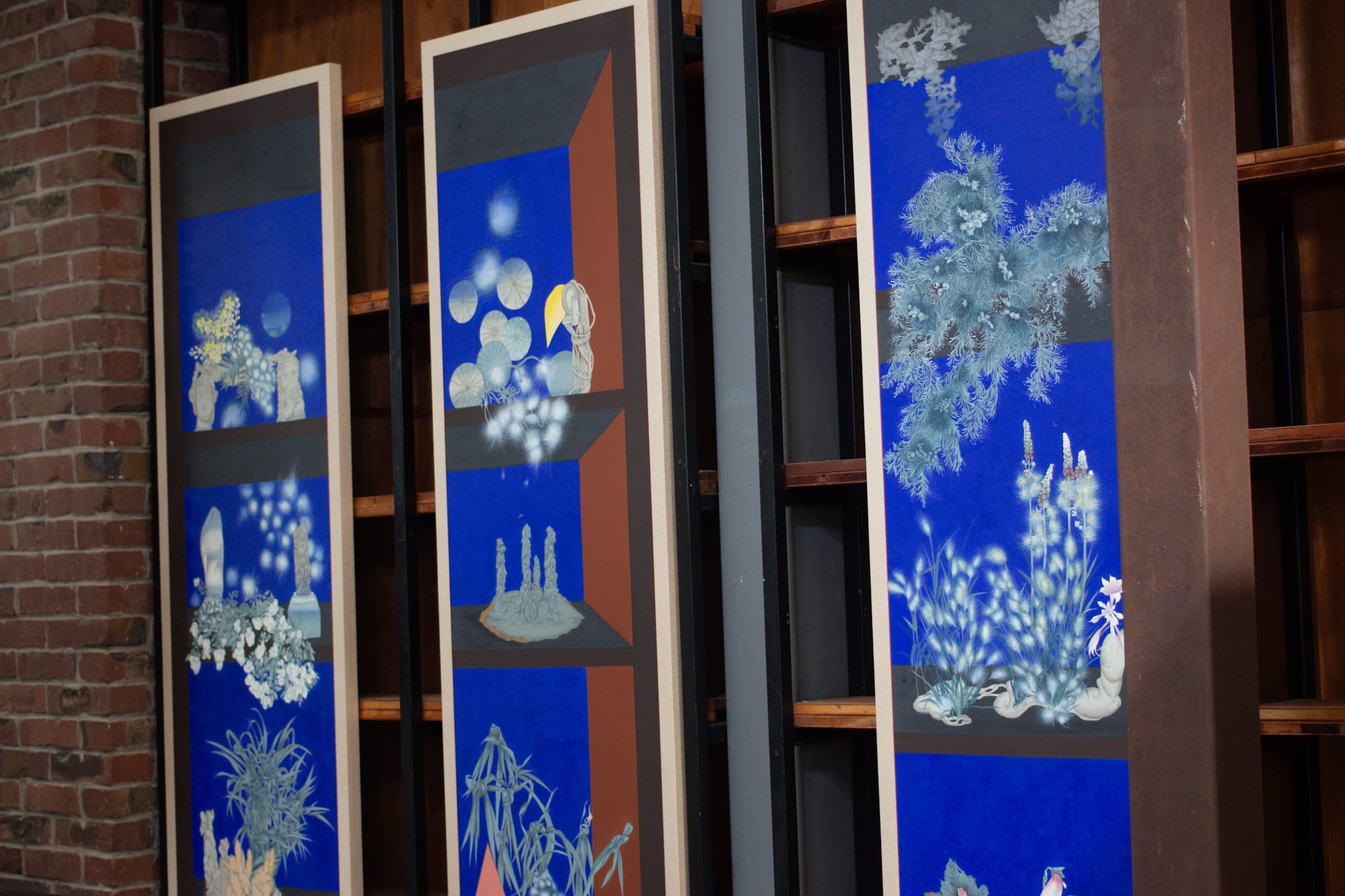 Three long paintings featuring a limited color palette. The paintings are of interior still life scenes on shelves with bright blue backgrounds. The objects are white and glowing.