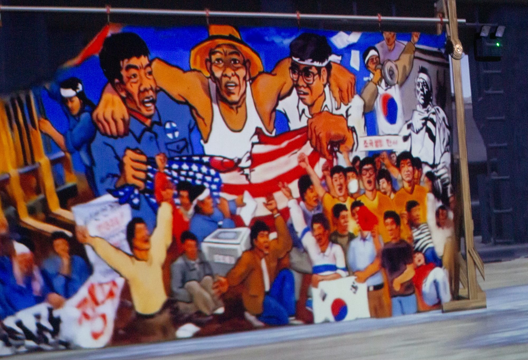 Large banner painting on wooden supports. Three figures stand with arms around each others' shoulders, ripping a U.S. flag. One is in a blue collared shirt, another is in a white tank top and farmer hat, and the last one is in a white collared shirt and white headband.