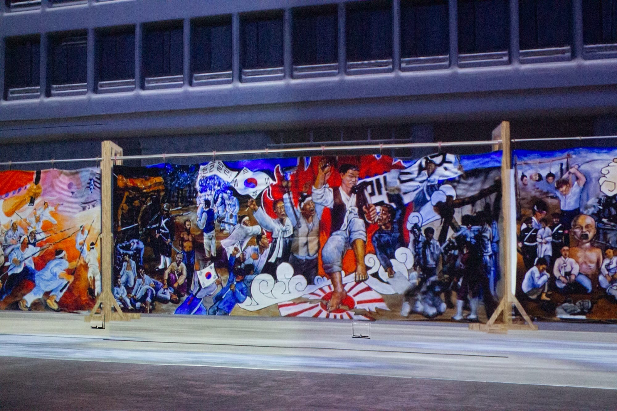 Three large panels of the banner painting. In the center, a figure steps on a red and white 'rising sun' flag. Behind are other activists, Korean flags, and signs.